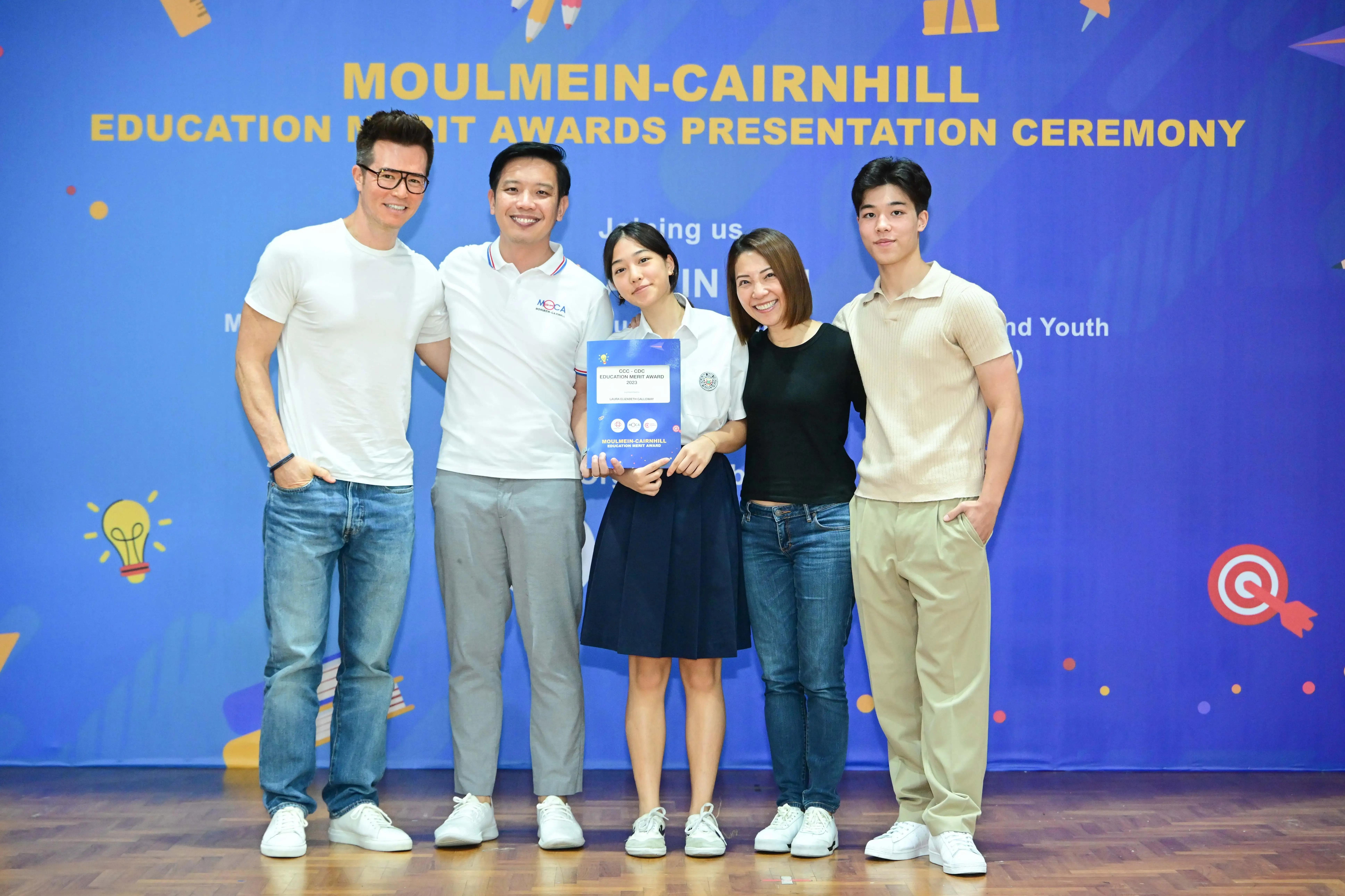 Five people pose smiling at the Moulmein-Cairnhill Education Merit Awards Presentation Ceremony, with a student holding an award.