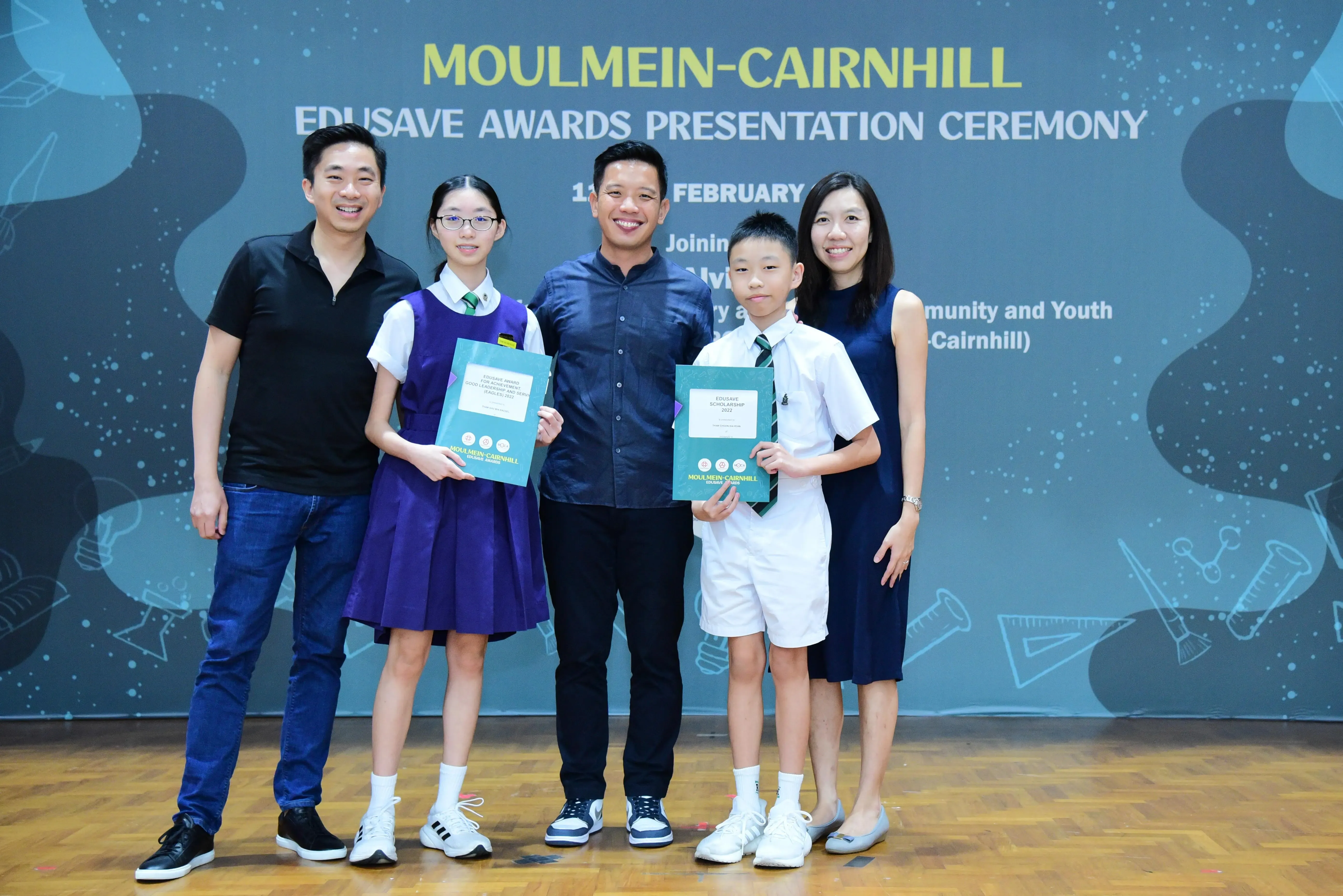 Five people pose at the Moulmein-Cairnhill Edusave Awards Presentation Ceremony, two children holding award certificates.