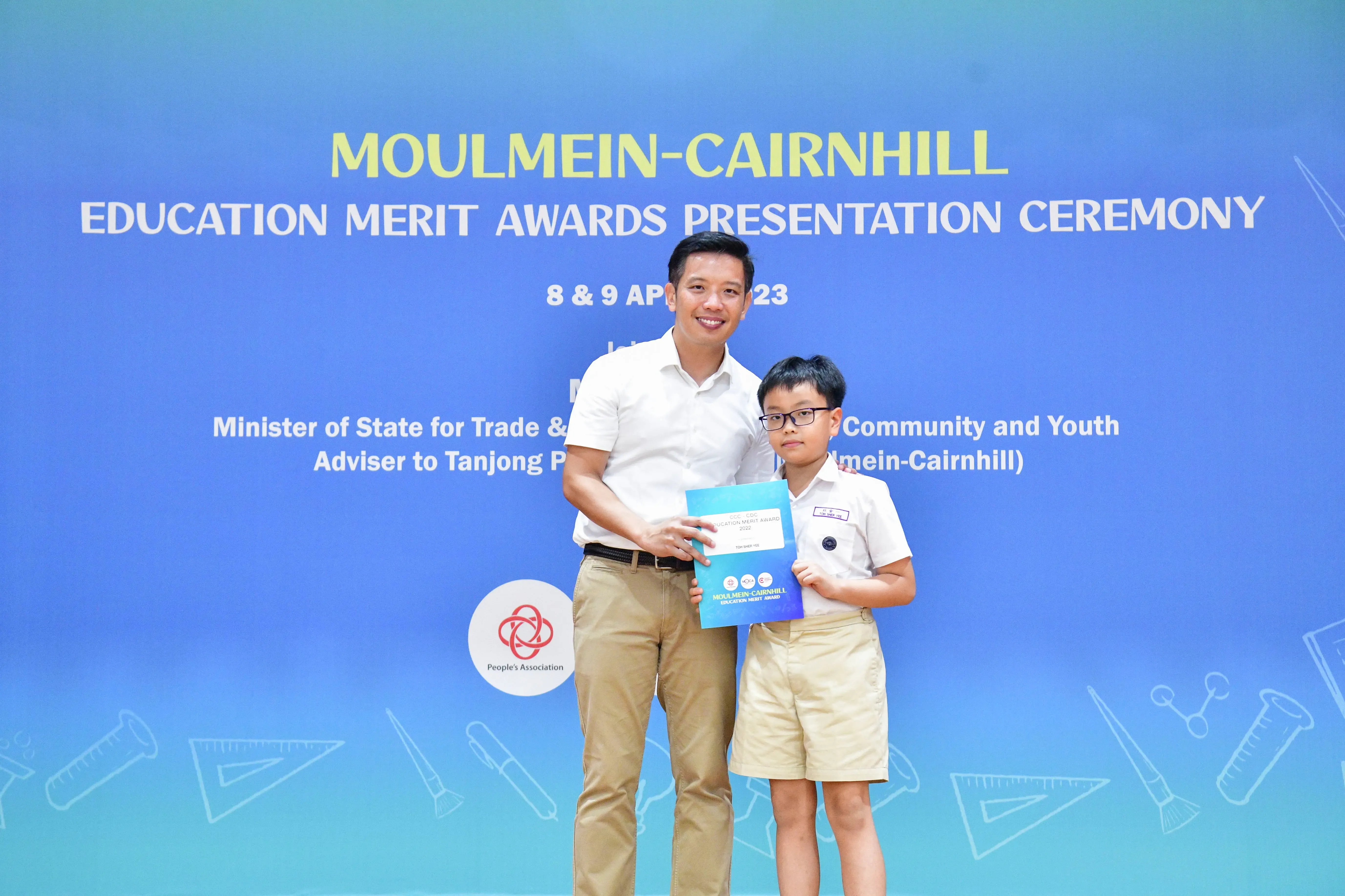 An adult man and a young boy in school uniform hold a certificate at the Moulmein-Cairnhill Education Merit Awards ceremony.