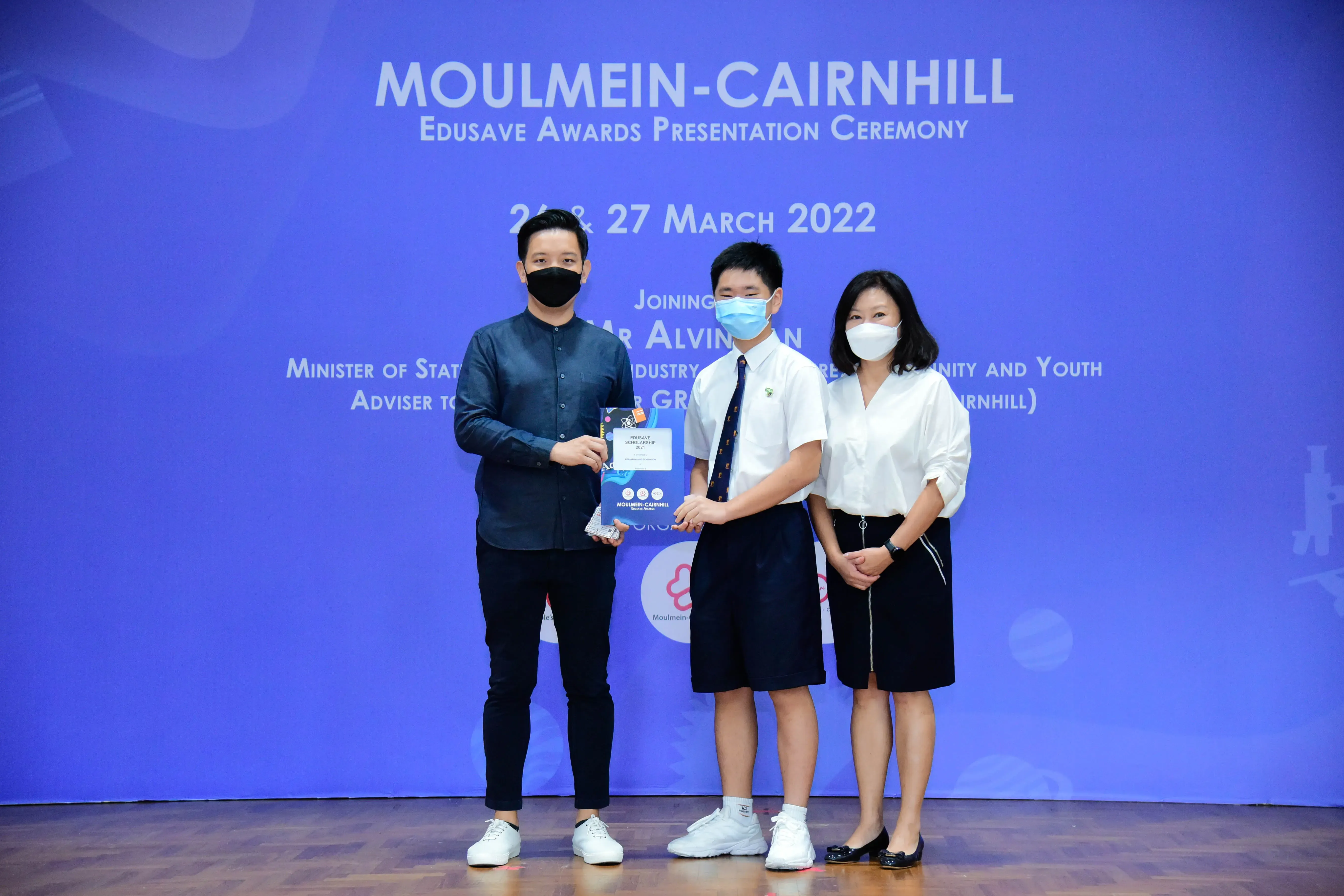 Three masked people pose on a stage at the Moulmein-Cairnhill Edusave Awards Presentation Ceremony, March 2022.