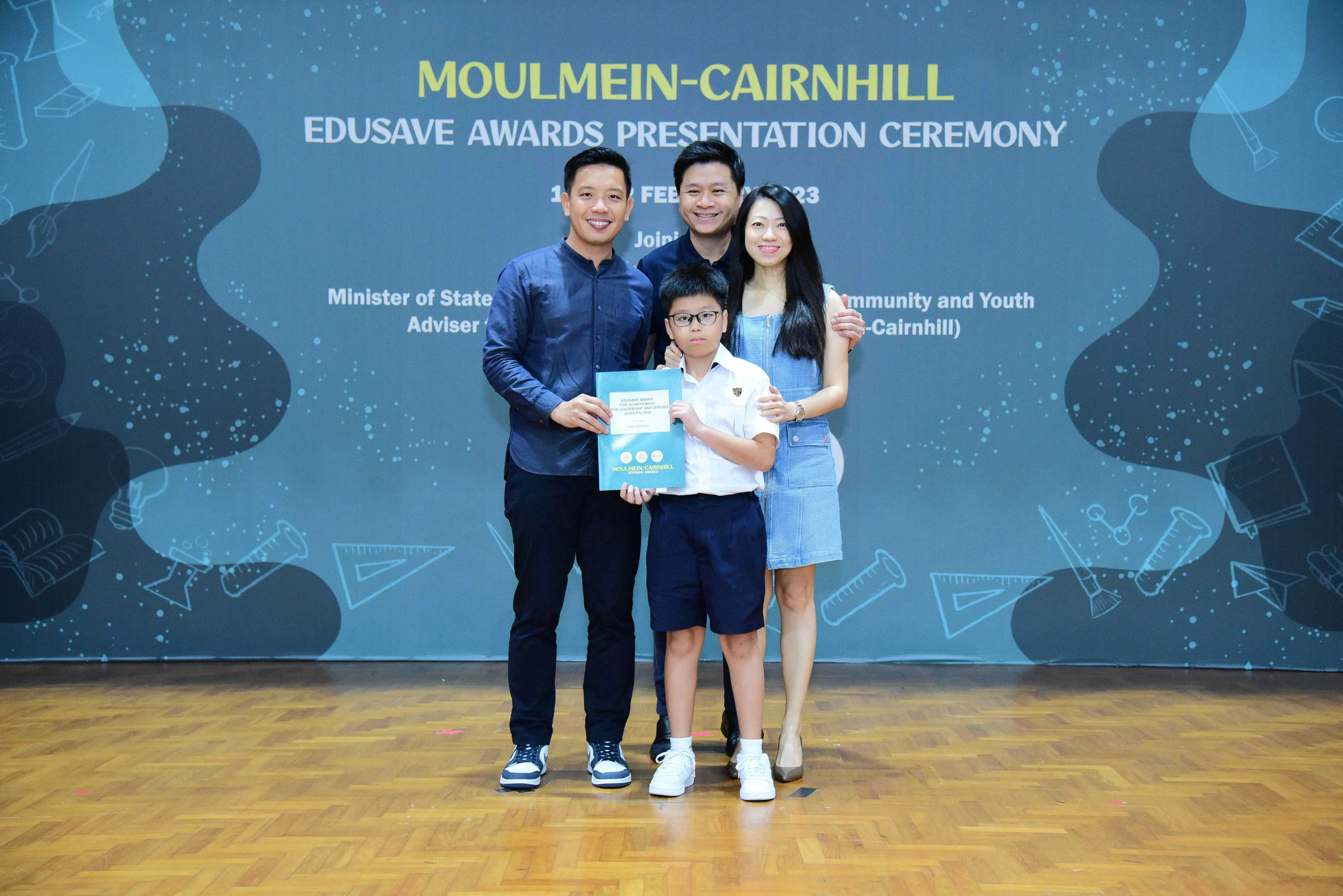 Four people pose at the Moulmein-Cairnhill Edusave Awards Presentation Ceremony, with a boy holding a certificate.