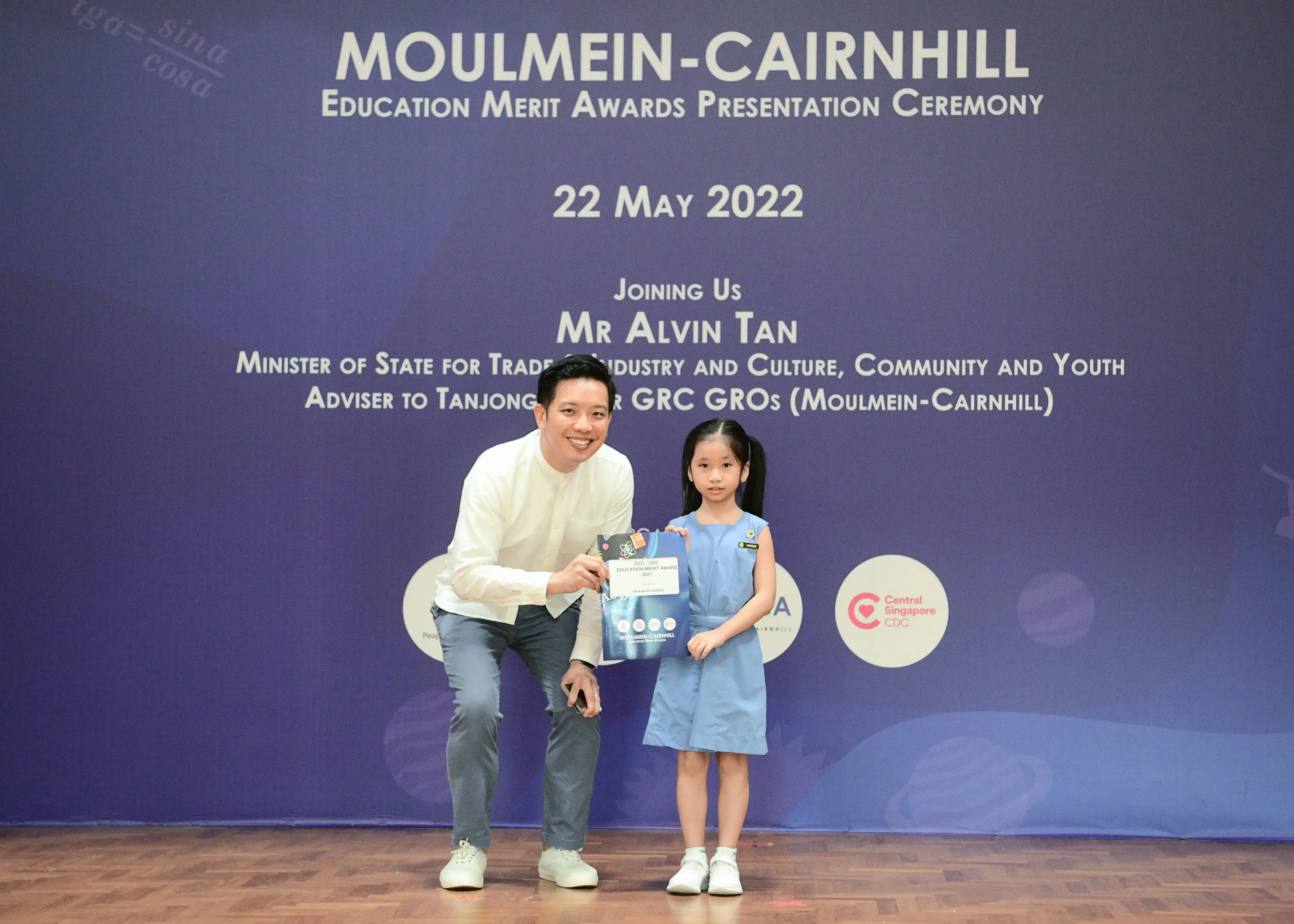 A man kneeling beside a young girl holding an award bag at the Moulmein-Cairnhill Education Merit Awards on 22 May 2022.