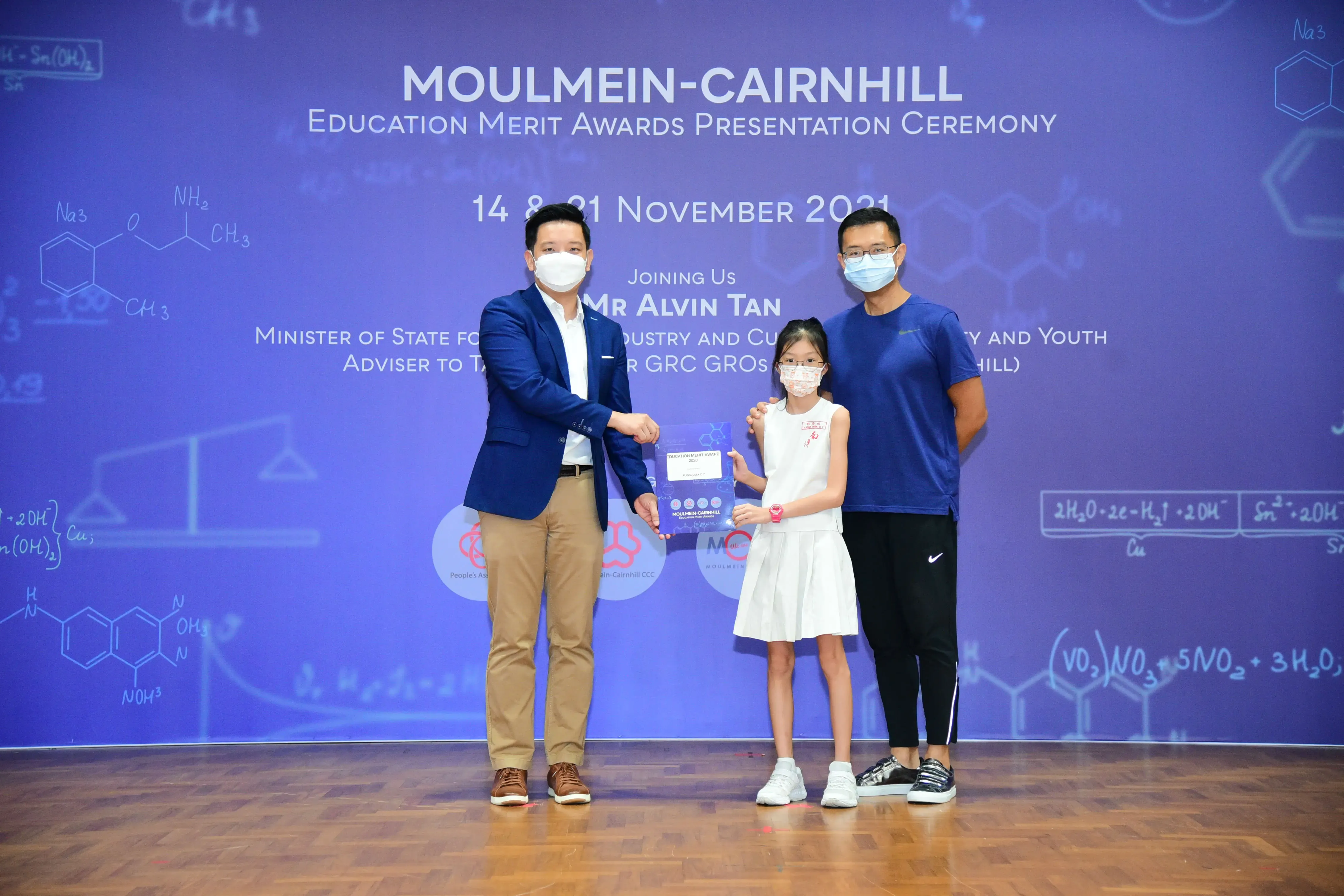 Two men and a young girl in masks pose on stage at the Moulmein-Cairnhill Education Merit Awards ceremony, November 2021.