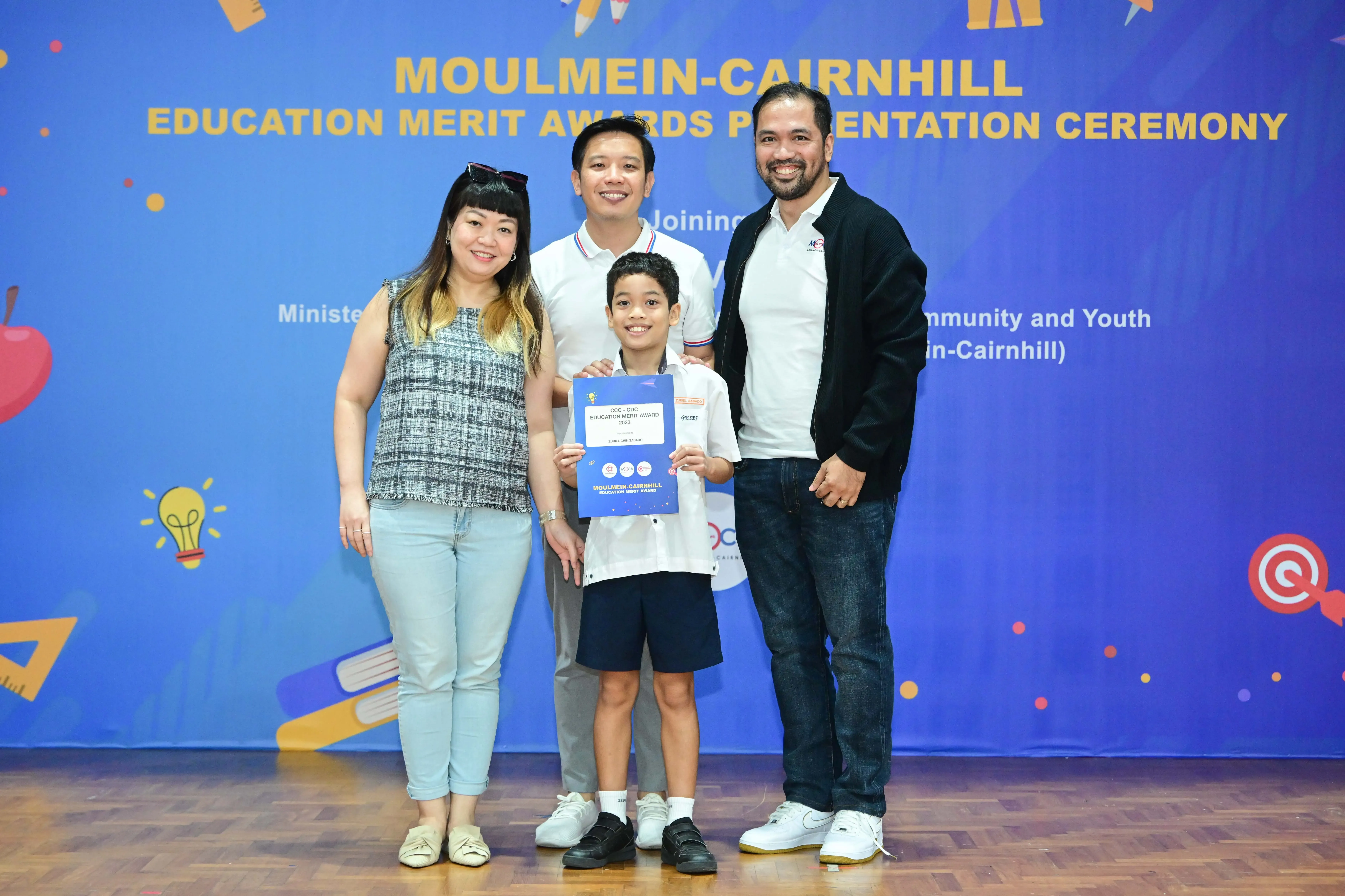 A boy holds an award certificate flanked by three adults at the Moulmein-Cairnhill Education Merit Awards ceremony.