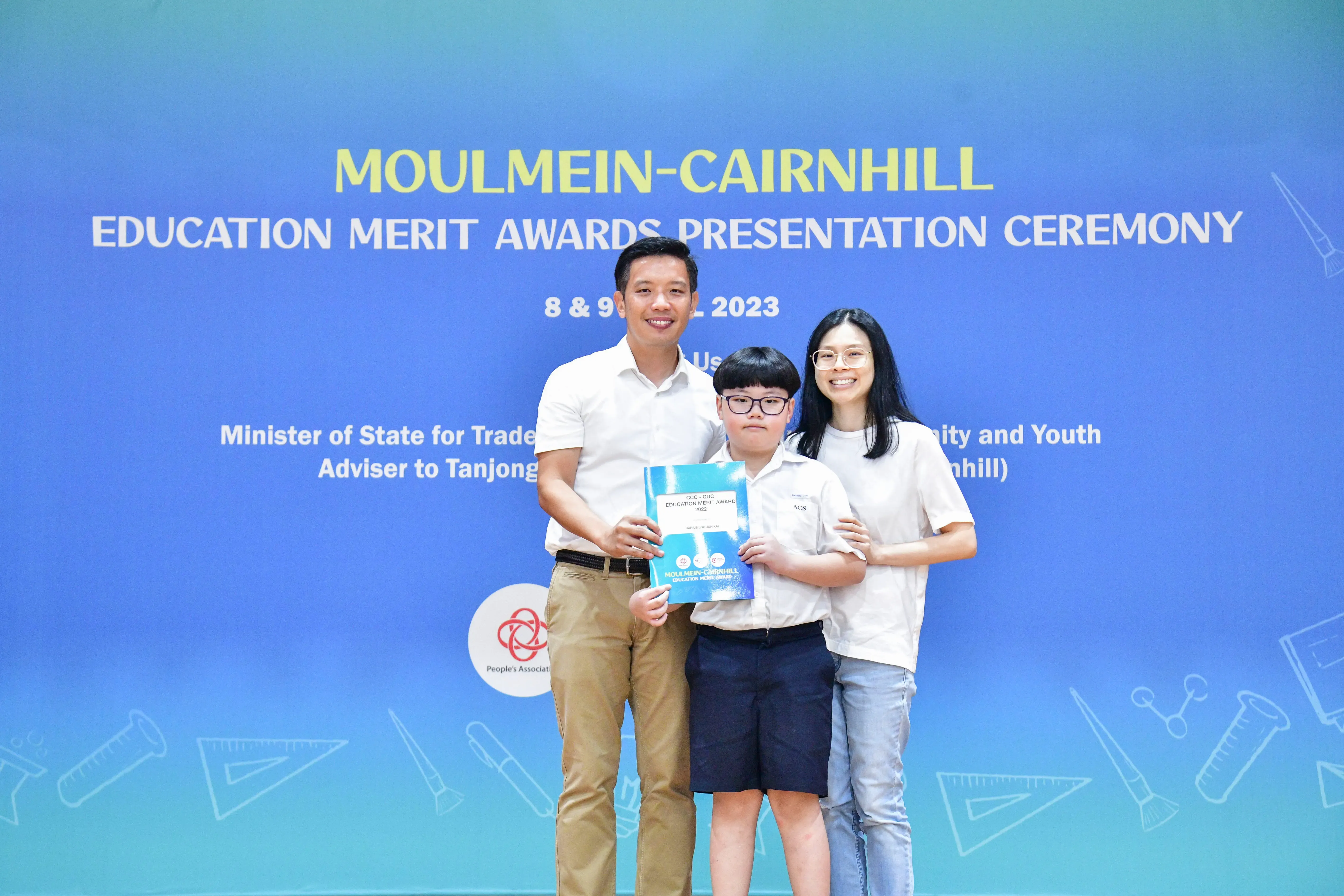 Two adults and a boy holding an award certificate pose before a Moulmein-Cairnhill Education Merit Awards backdrop.