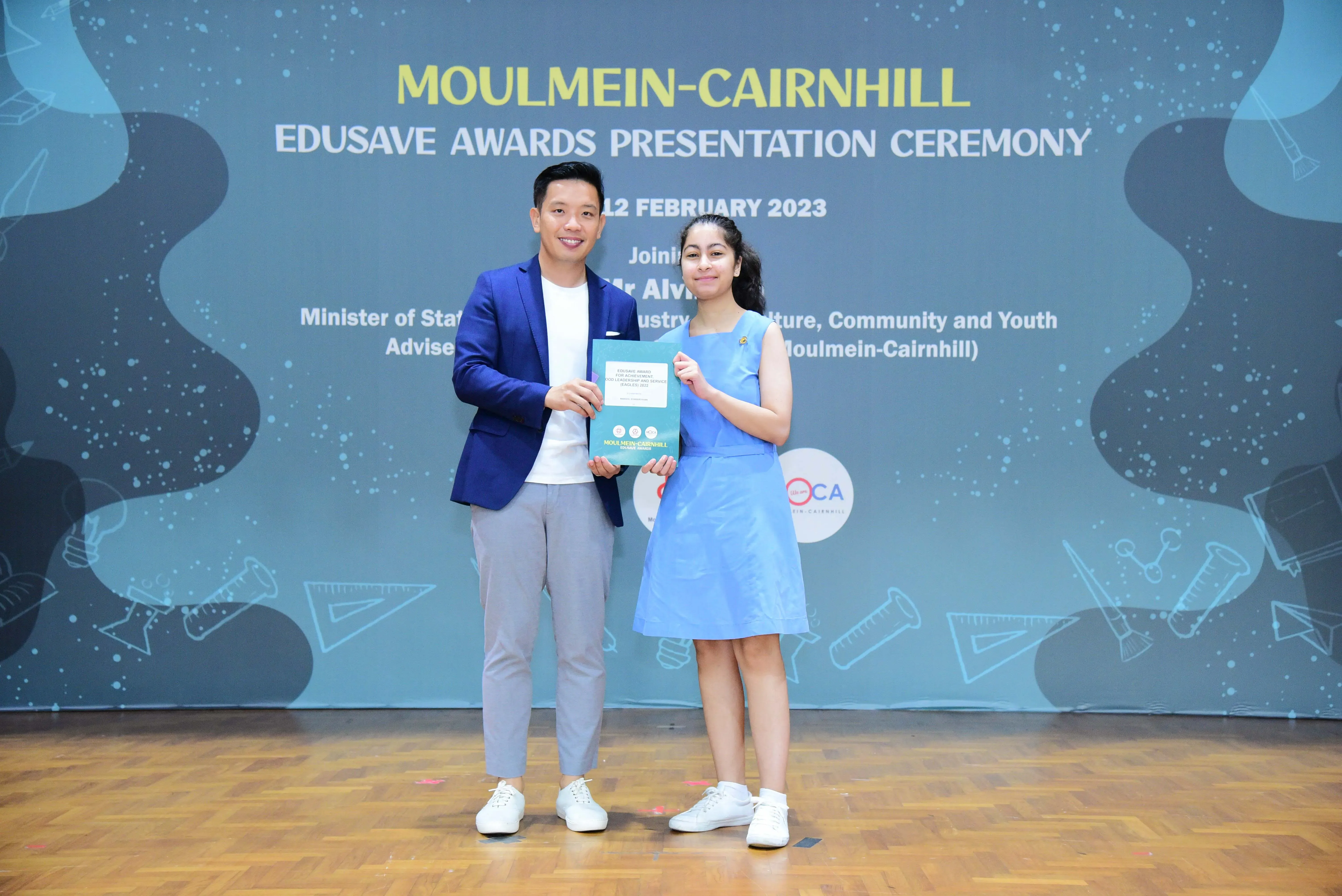 A man in a blue blazer and a girl in a blue dress hold a certificate at the Moulmein-Cairnhill Edusave Awards Ceremony 2023.