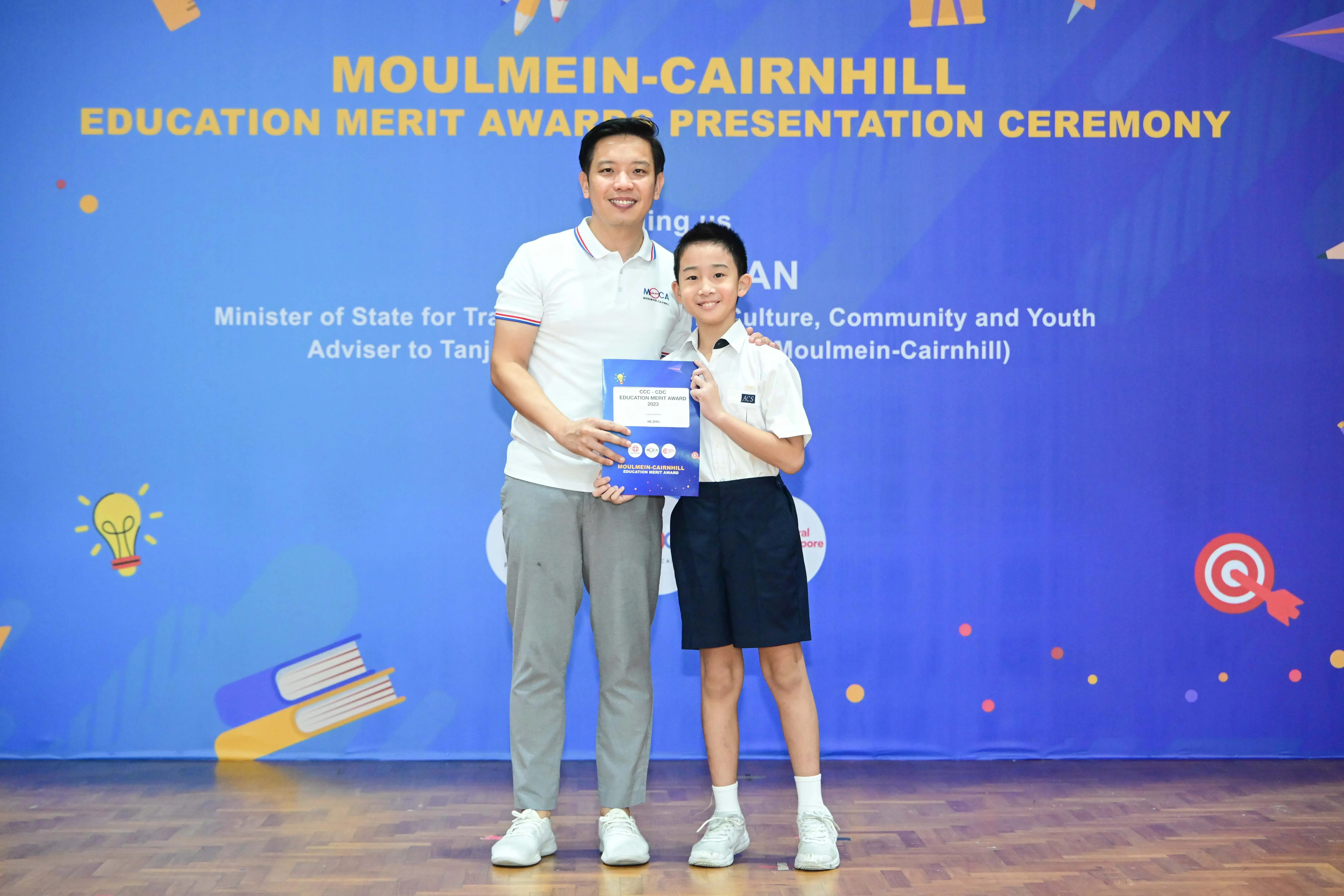 Adult and young boy in school uniform hold an award certificate at Moulmein-Cairnhill Education Merit Awards ceremony.