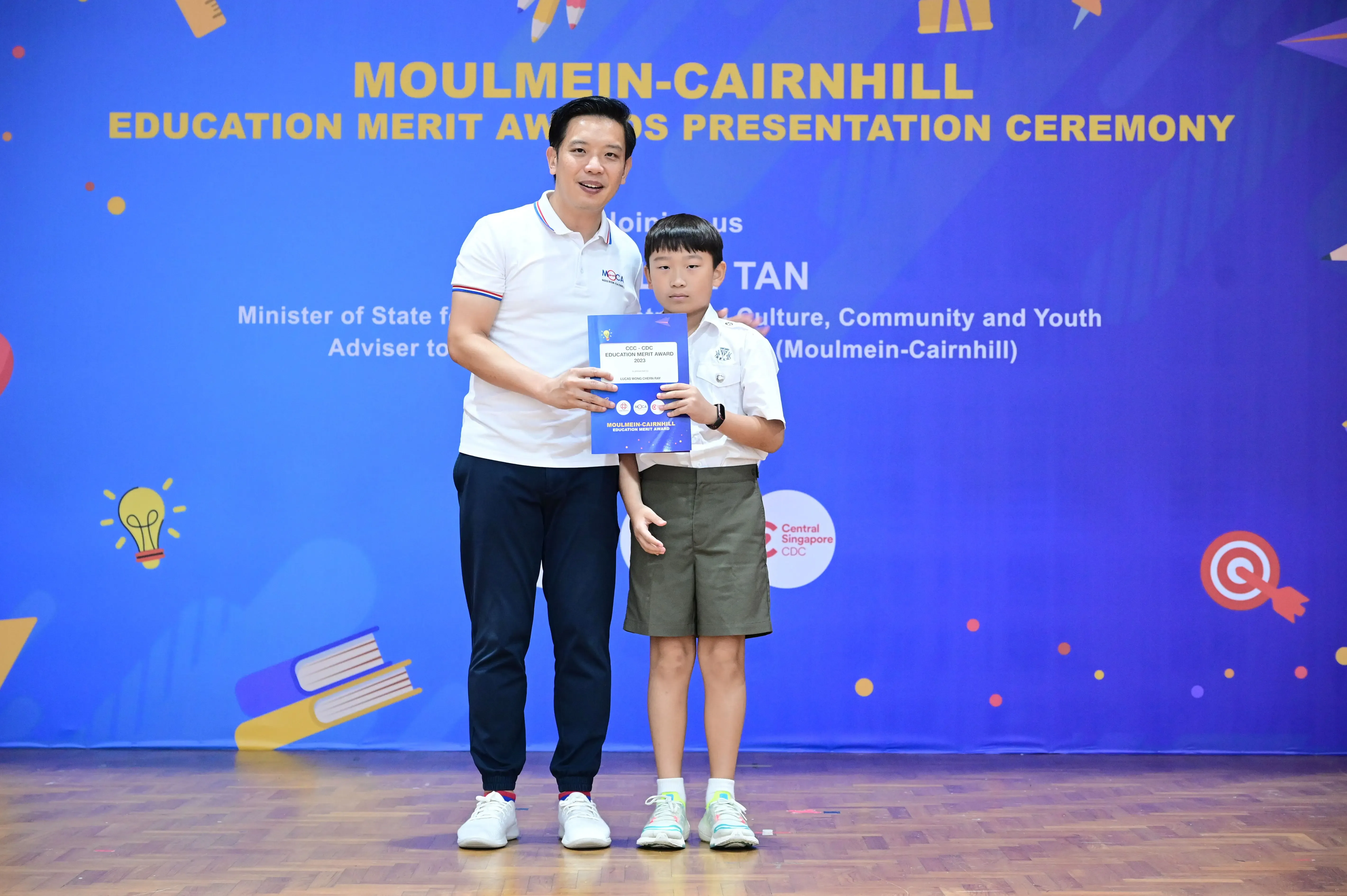 An adult and a young student hold an award certificate at the Moulmein-Cairnhill Education Merit Awards Presentation Ceremony.