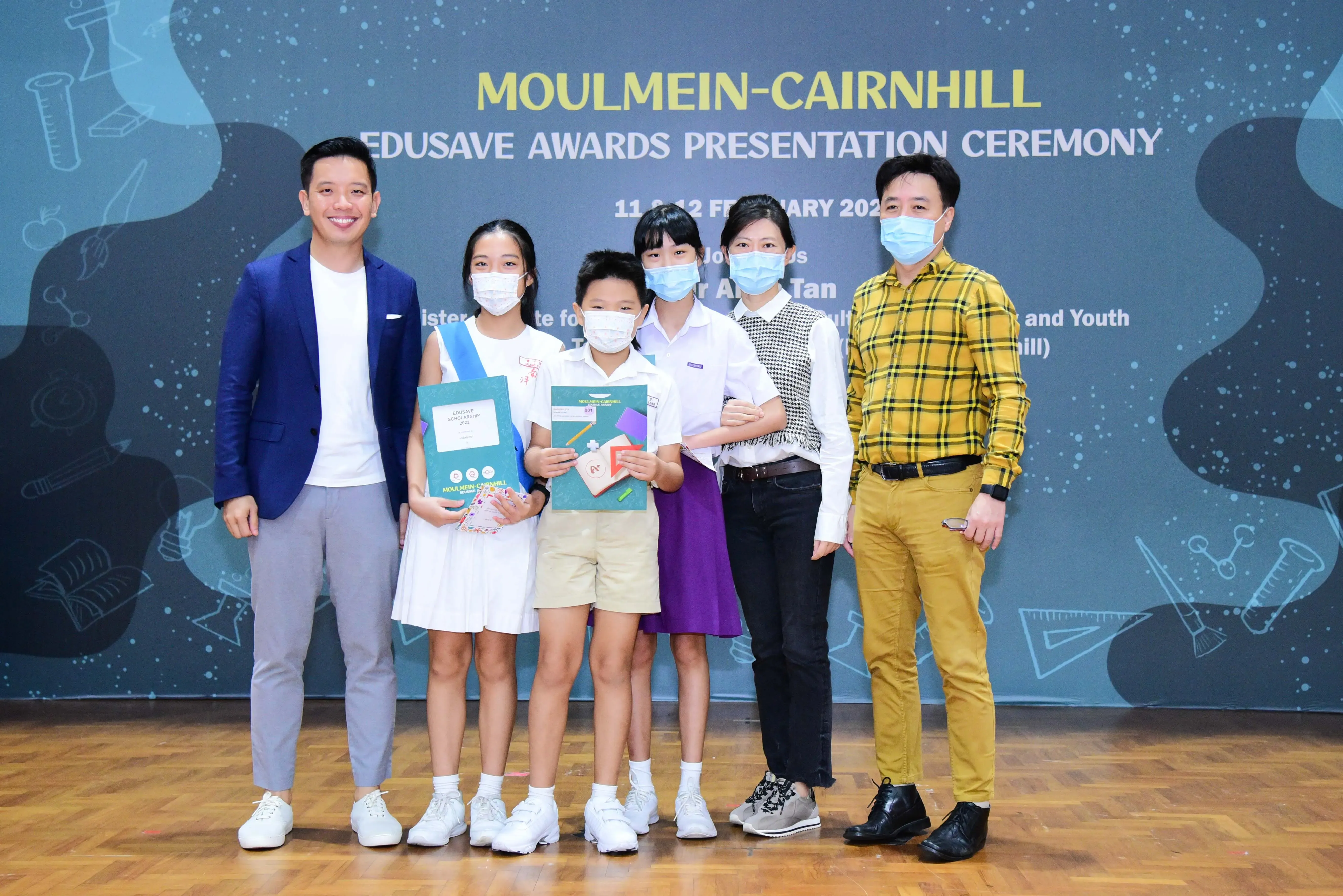 Six people wearing masks pose at the Moulmein-Cairnhill Edusave Awards Presentation Ceremony, with students holding award folders.