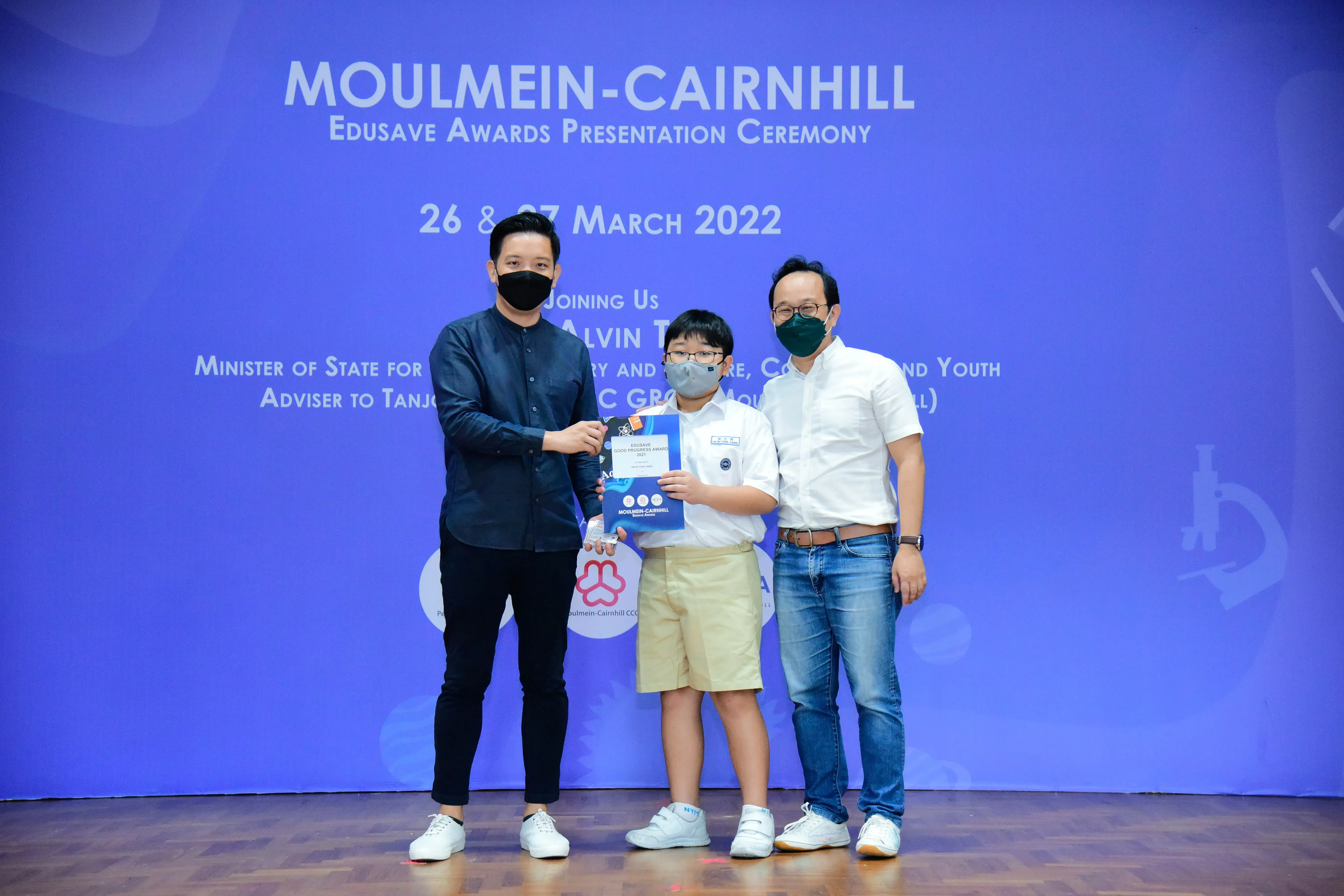 Three masked people pose at the Moulmein-Cairnhill Edusave Awards Ceremony on 26-27 March 2022, holding an award.