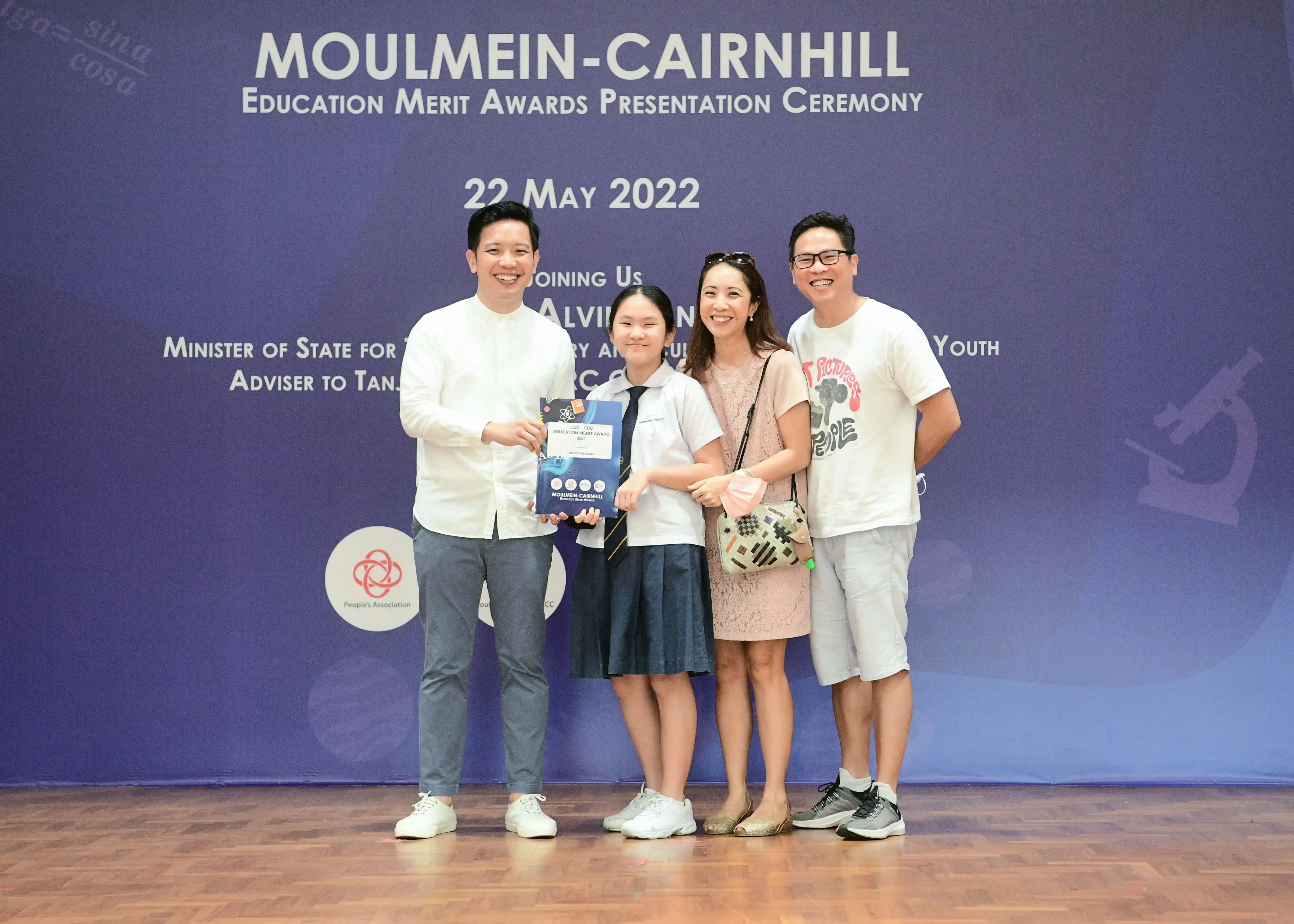 Four people pose smiling before a Moulmein-Cairnhill Education Merit Awards Presentation Ceremony banner dated 22 May 2022.