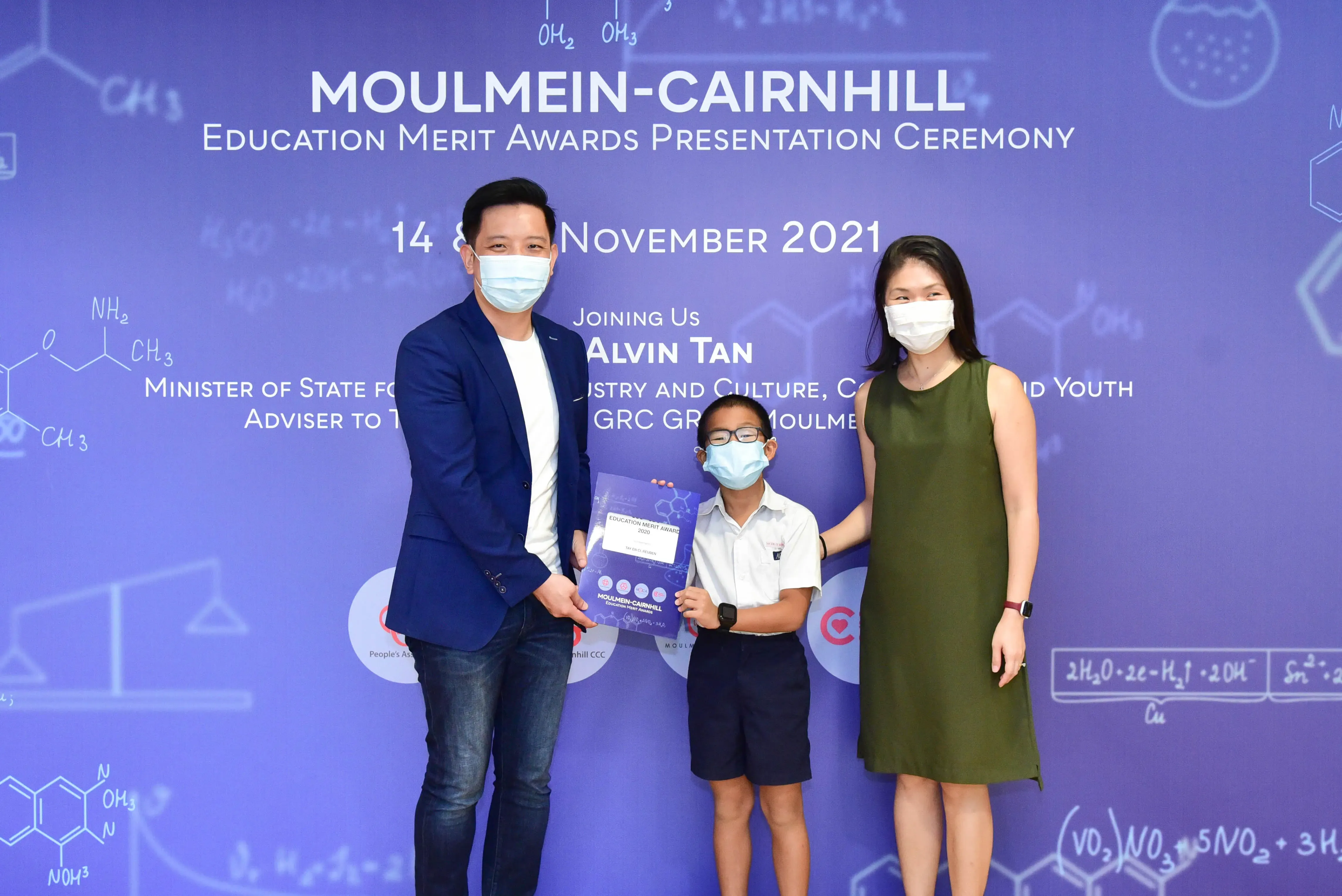 Two masked adults and a masked child hold an award certificate at the Moulmein-Cairnhill Education Merit Awards 2021.