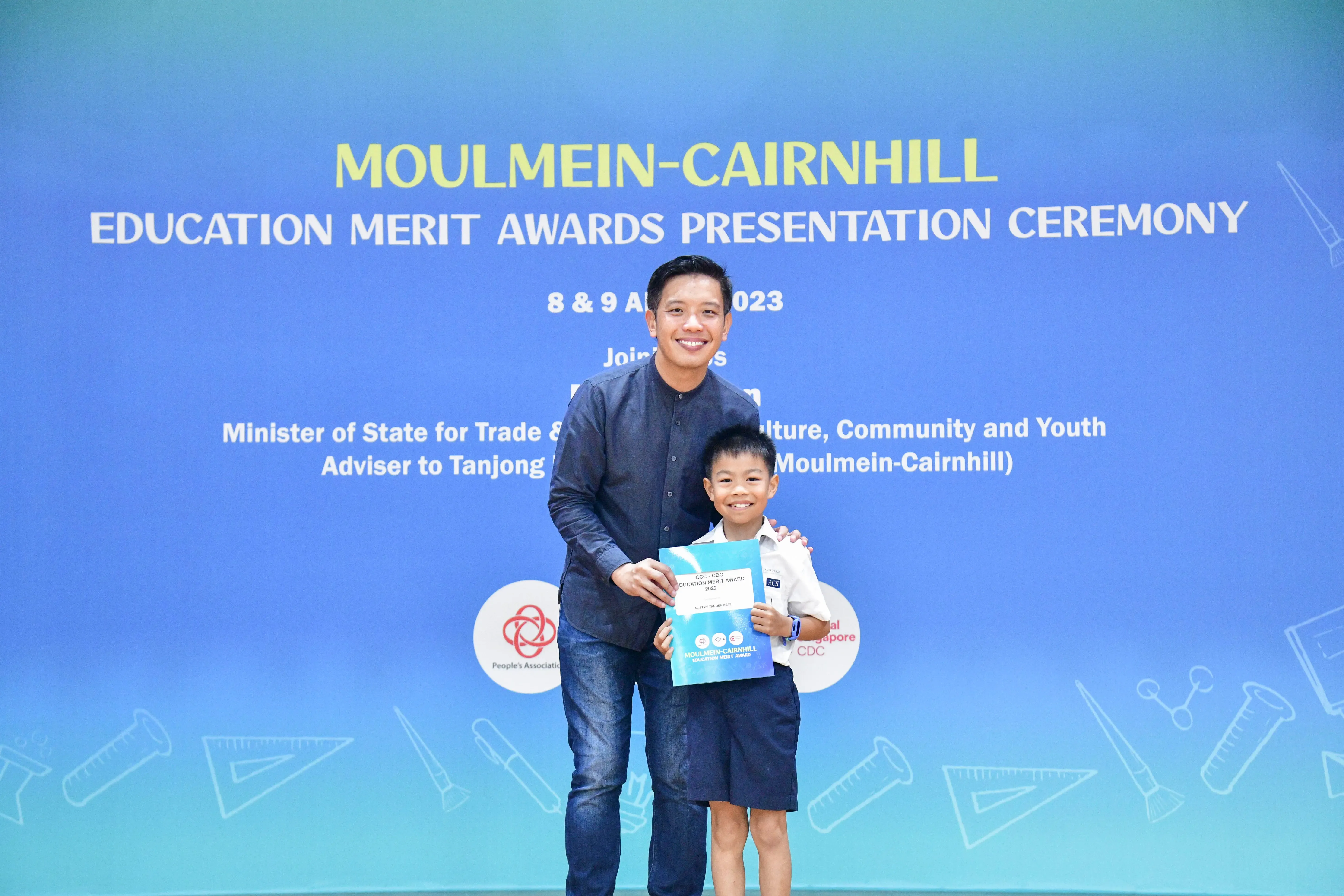 A man and a young boy holding an award certificate stand before a blue Moulmein-Cairnhill Education Merit Awards banner.