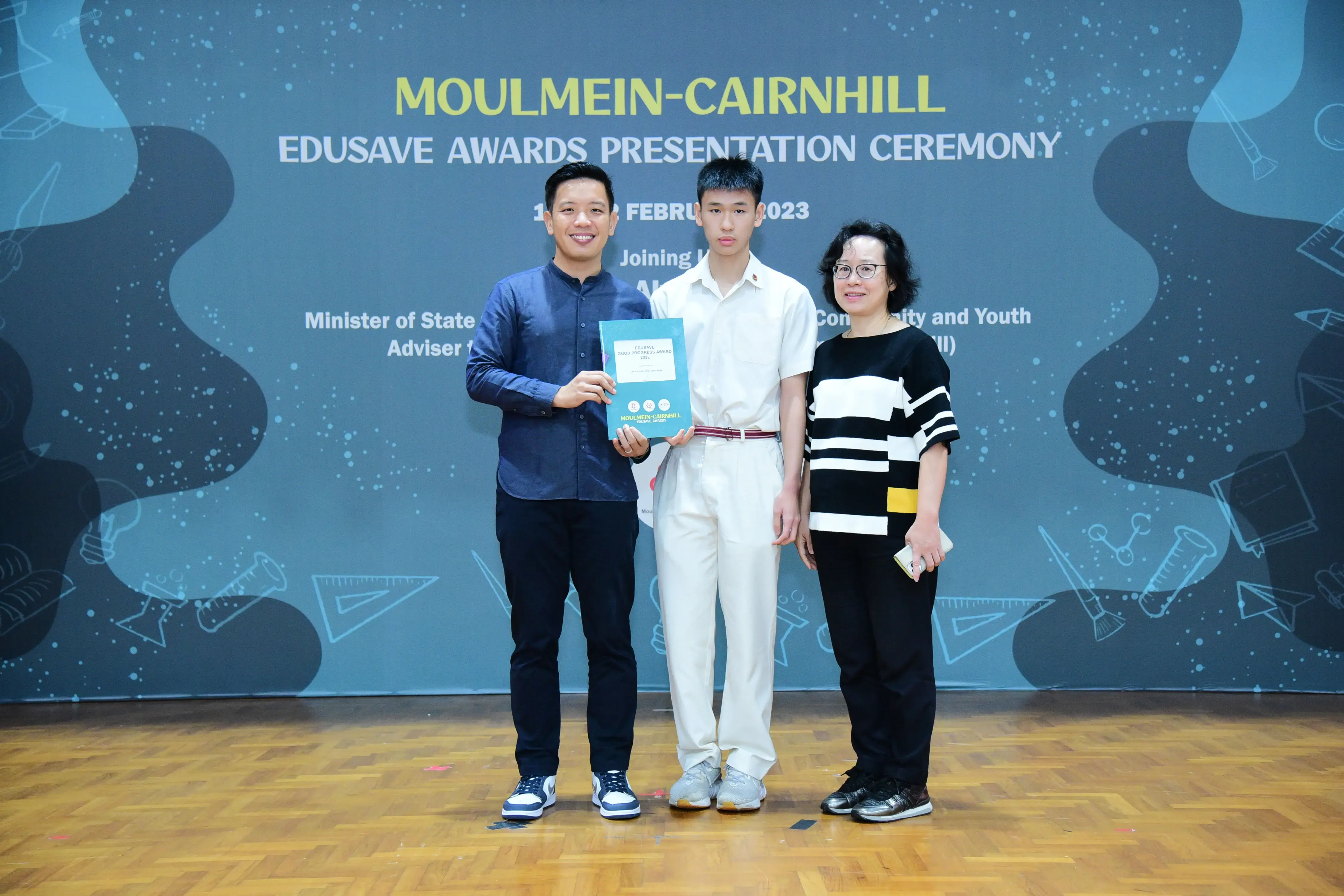 Three people stand on a stage holding an award booklet at the Moulmein-Cairnhill Edusave Awards Ceremony 2023.