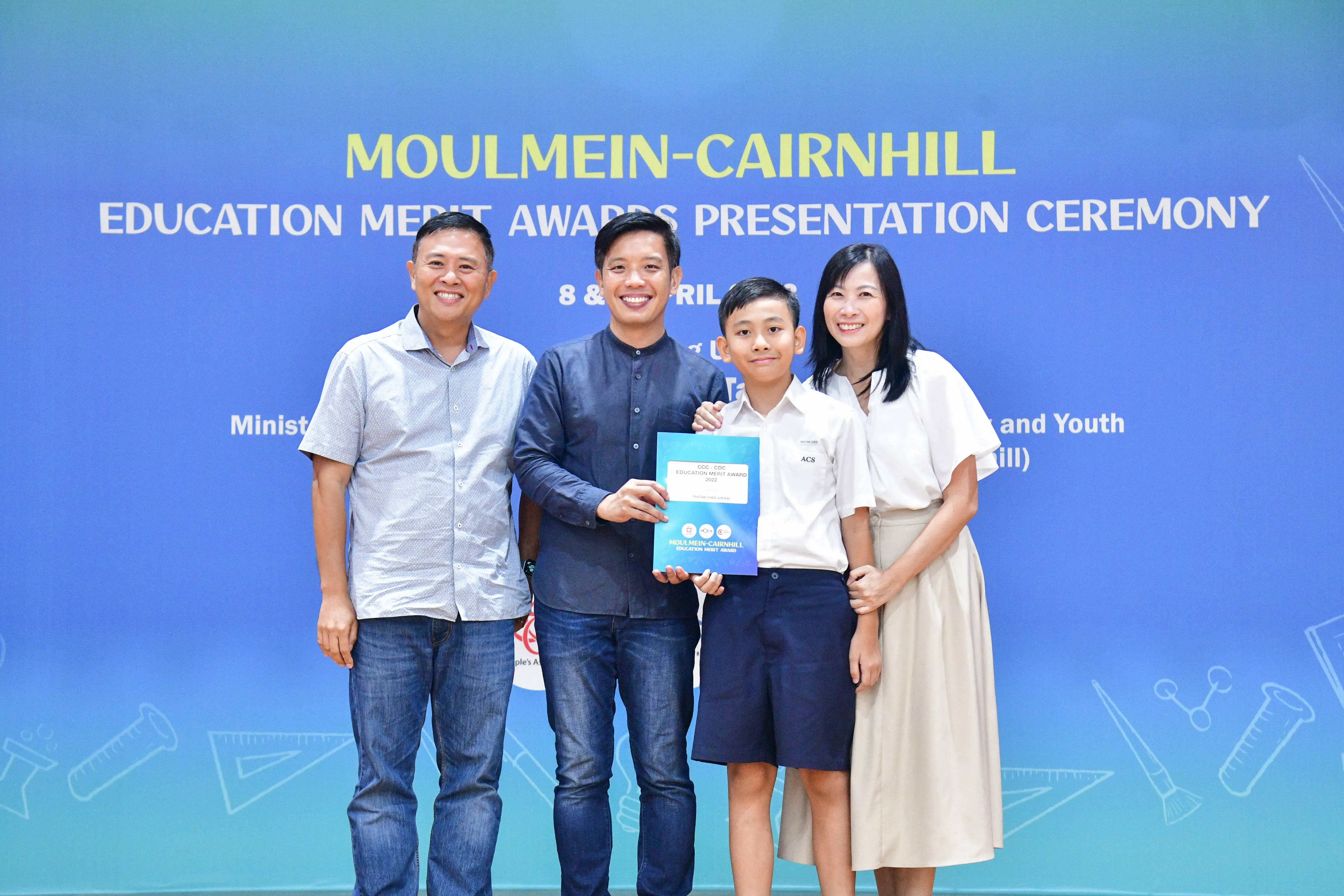 Four people pose at the Moulmein-Cairnhill Education Merit Awards Presentation Ceremony, with a boy holding an award certificate.