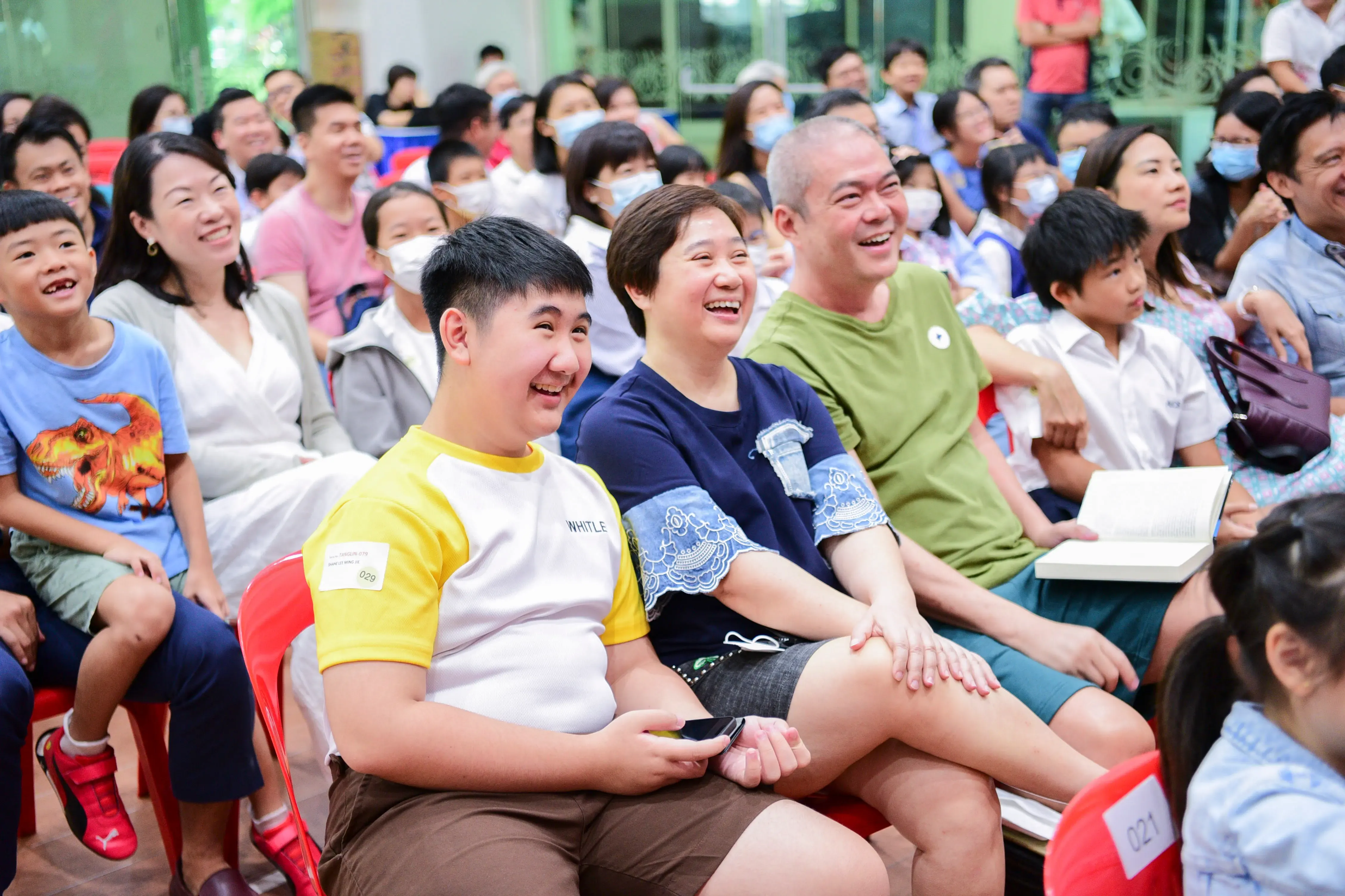 A large crowd of smiling adults and children sit in chairs at an indoor gathering, with three people laughing in the foreground.