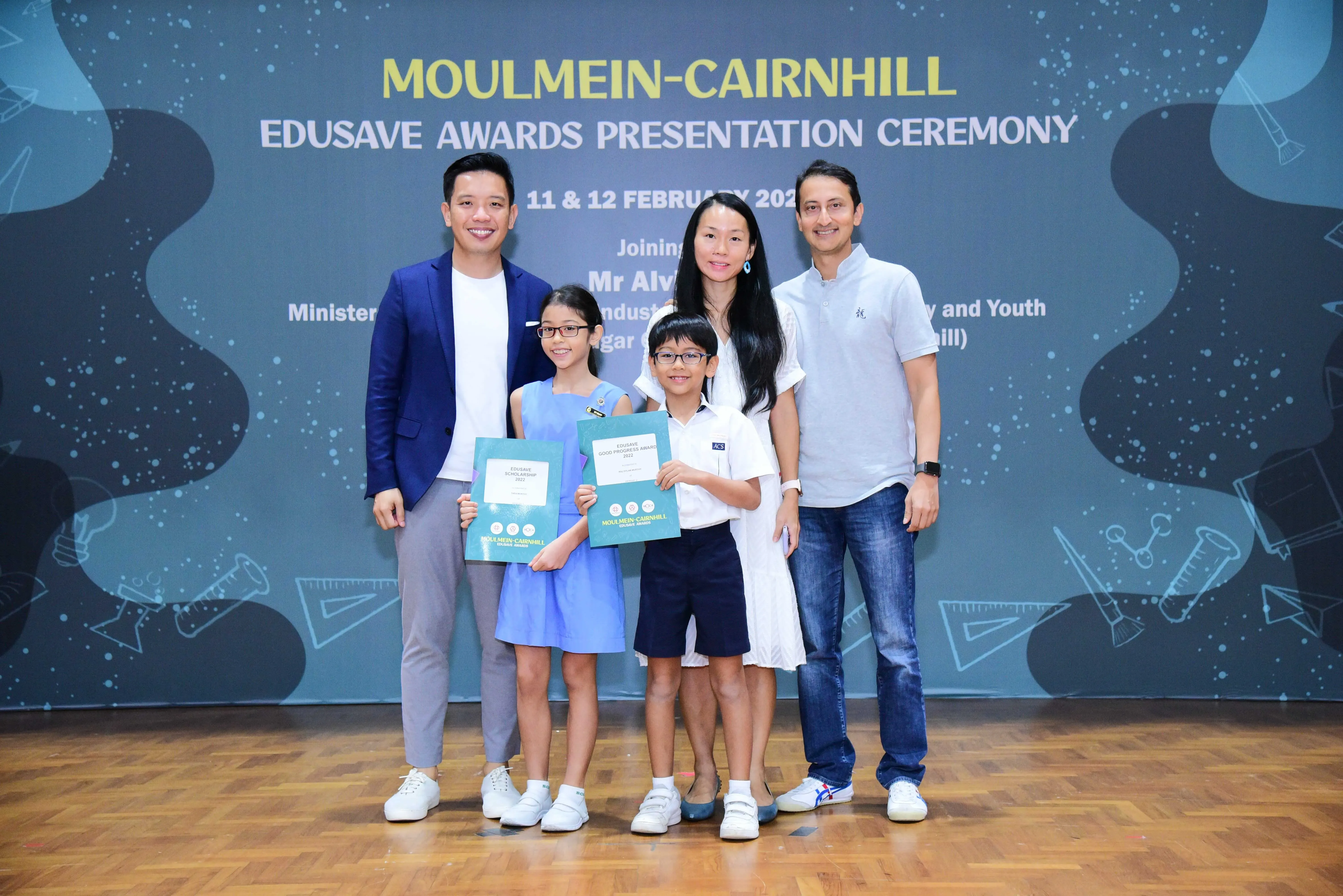 Two adults and two children hold award certificates in front of a Moulmein-Cairnhill Edusave Awards Ceremony banner.
