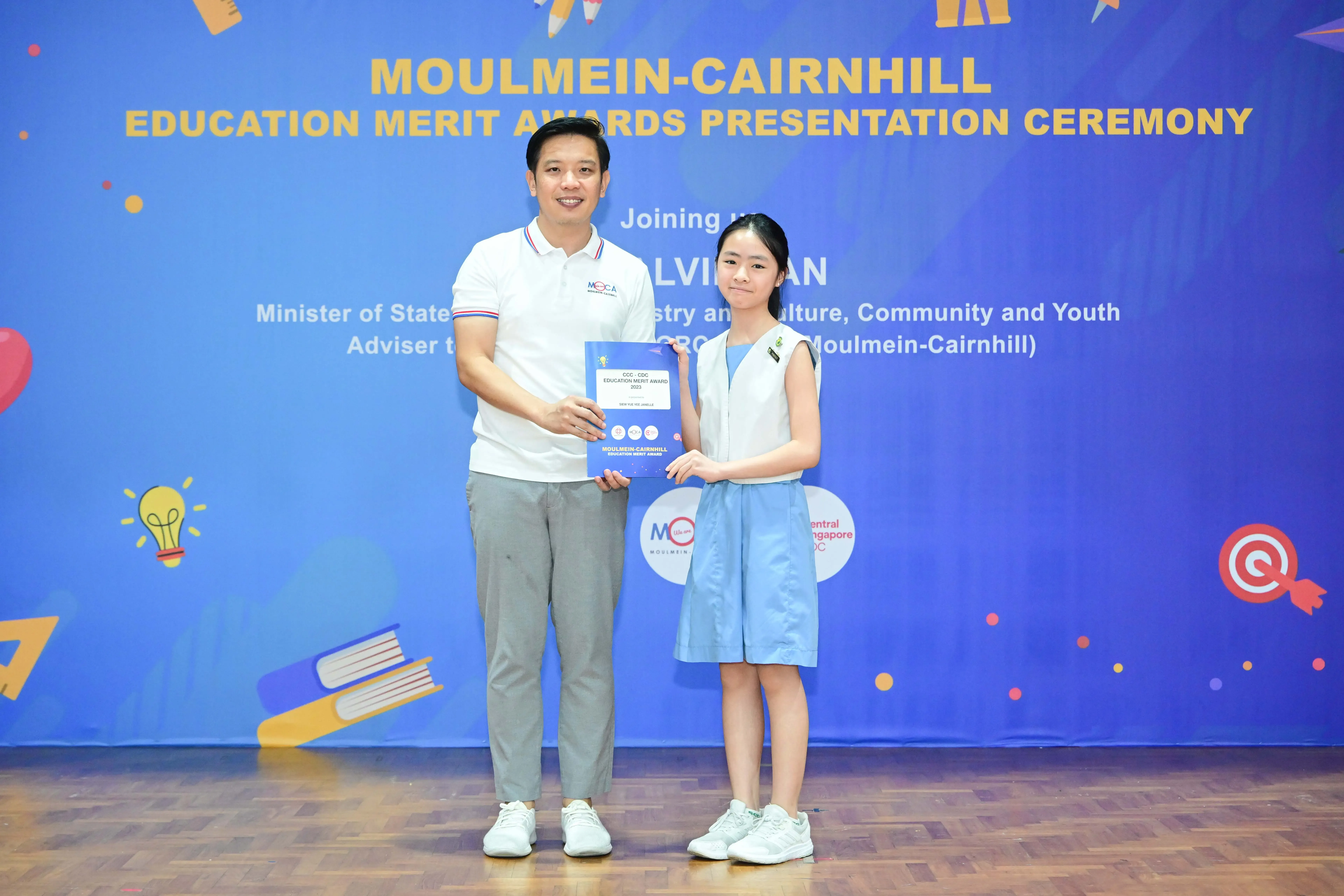A man and a young girl hold a blue award certificate at the Moulmein-Cairnhill Education Merit Awards ceremony.