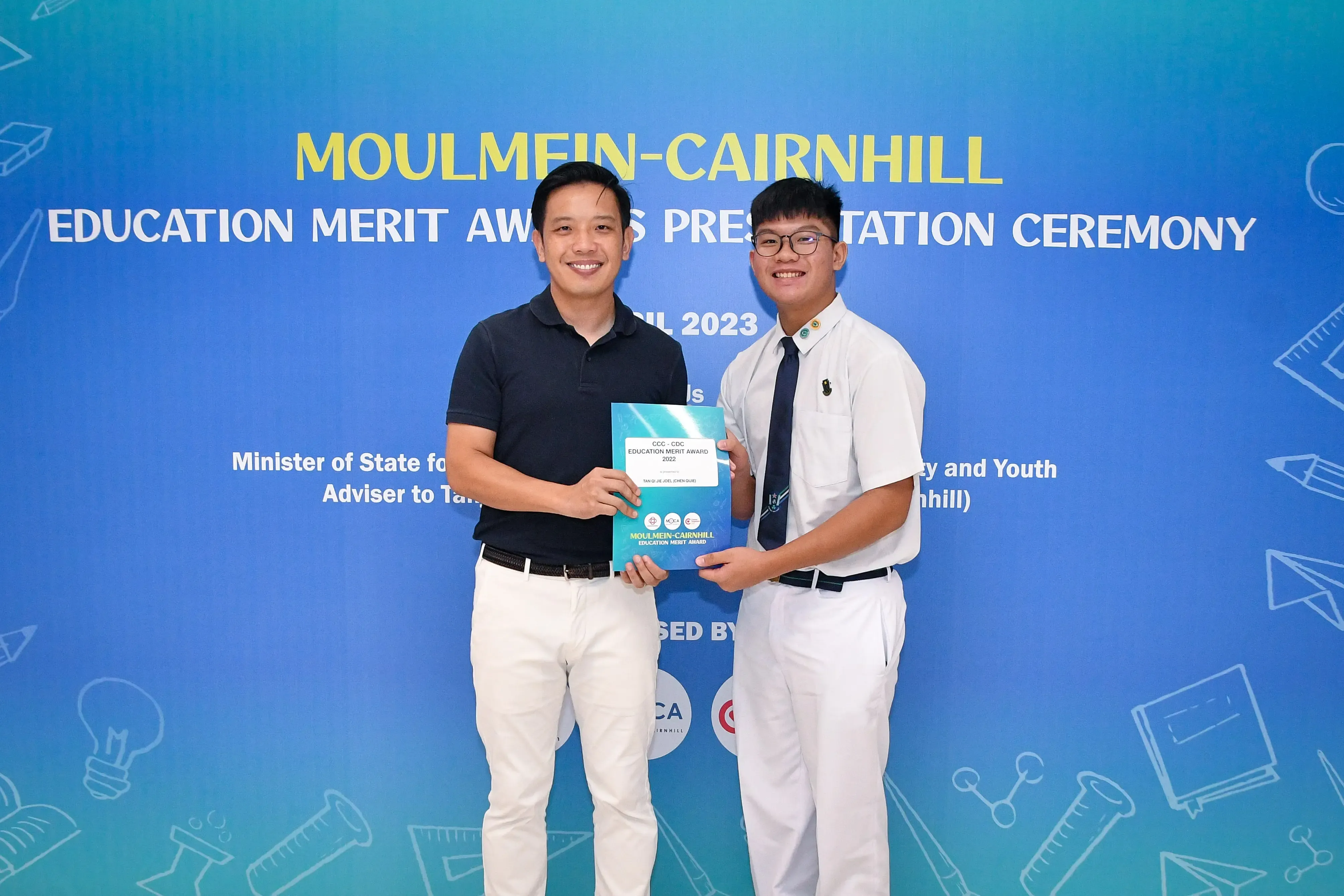 Two smiling men hold a teal award booklet at the Moulmein-Cairnhill Education Merit Awards Presentation Ceremony 2023.