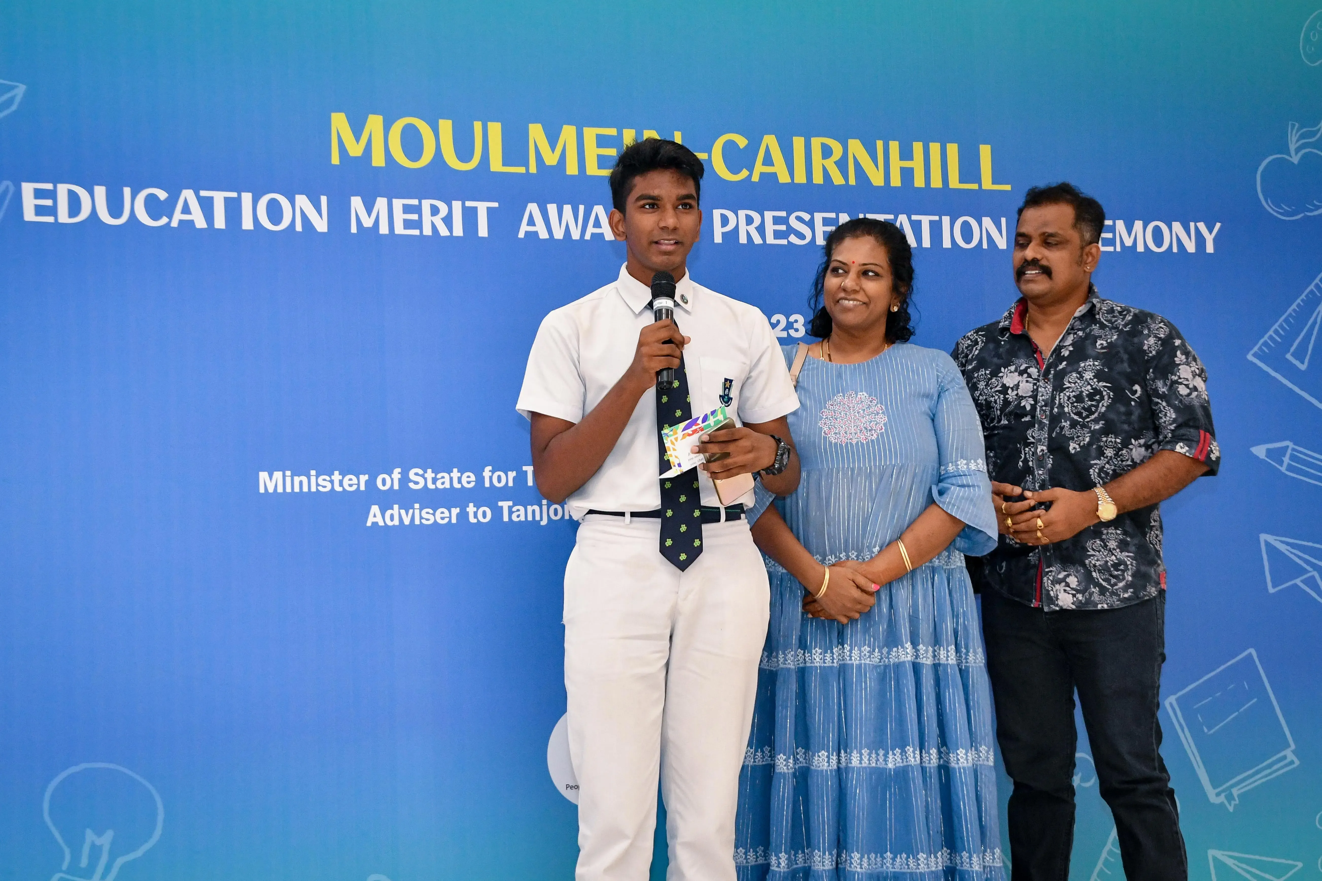 A young male student in white uniform speaks into a microphone at the Moulmein-Cairnhill Education Merit Award ceremony with two adults beside him.