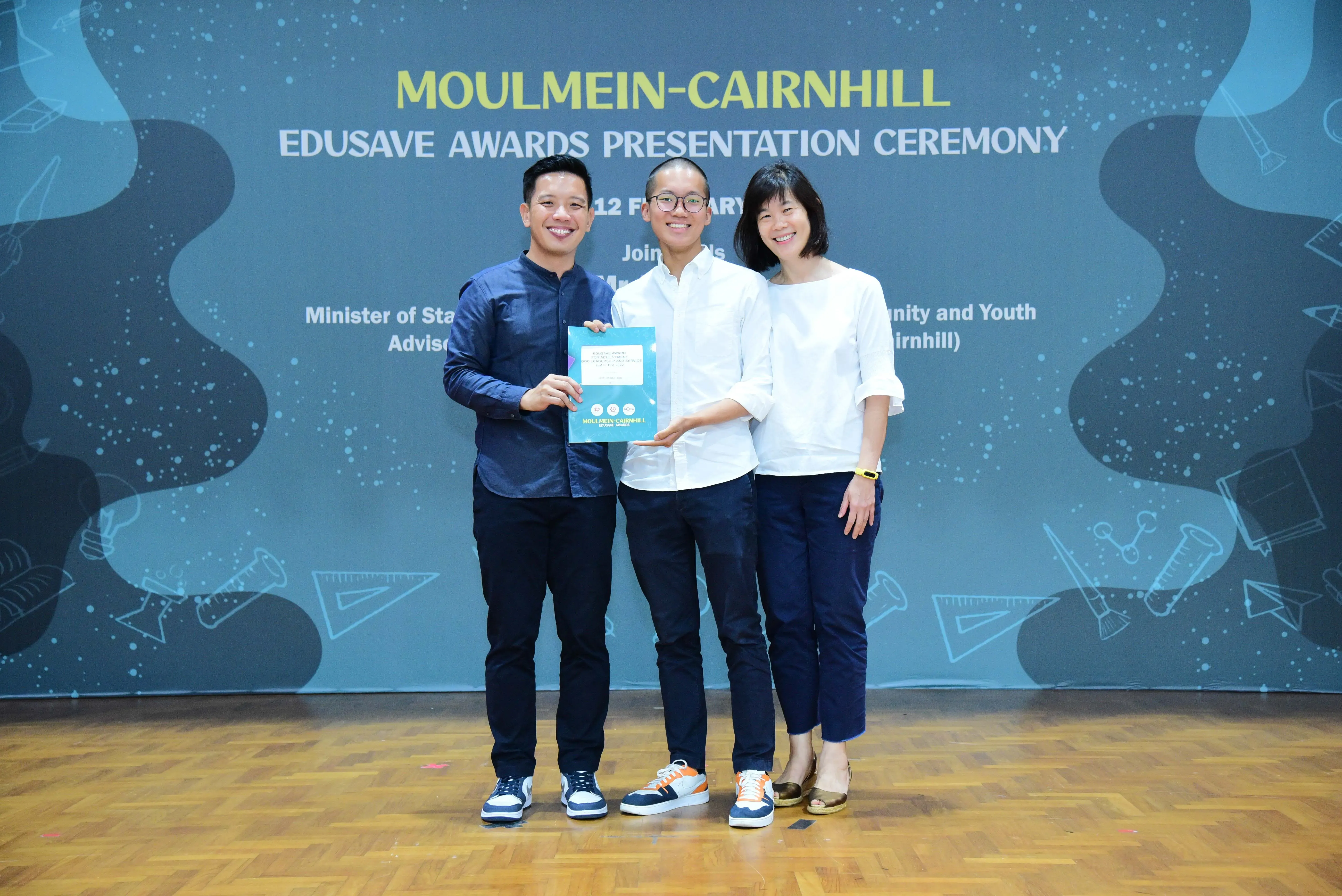 Three smiling people hold an award booklet in front of a Moulmein-Cairnhill Edusave Awards Presentation Ceremony banner.