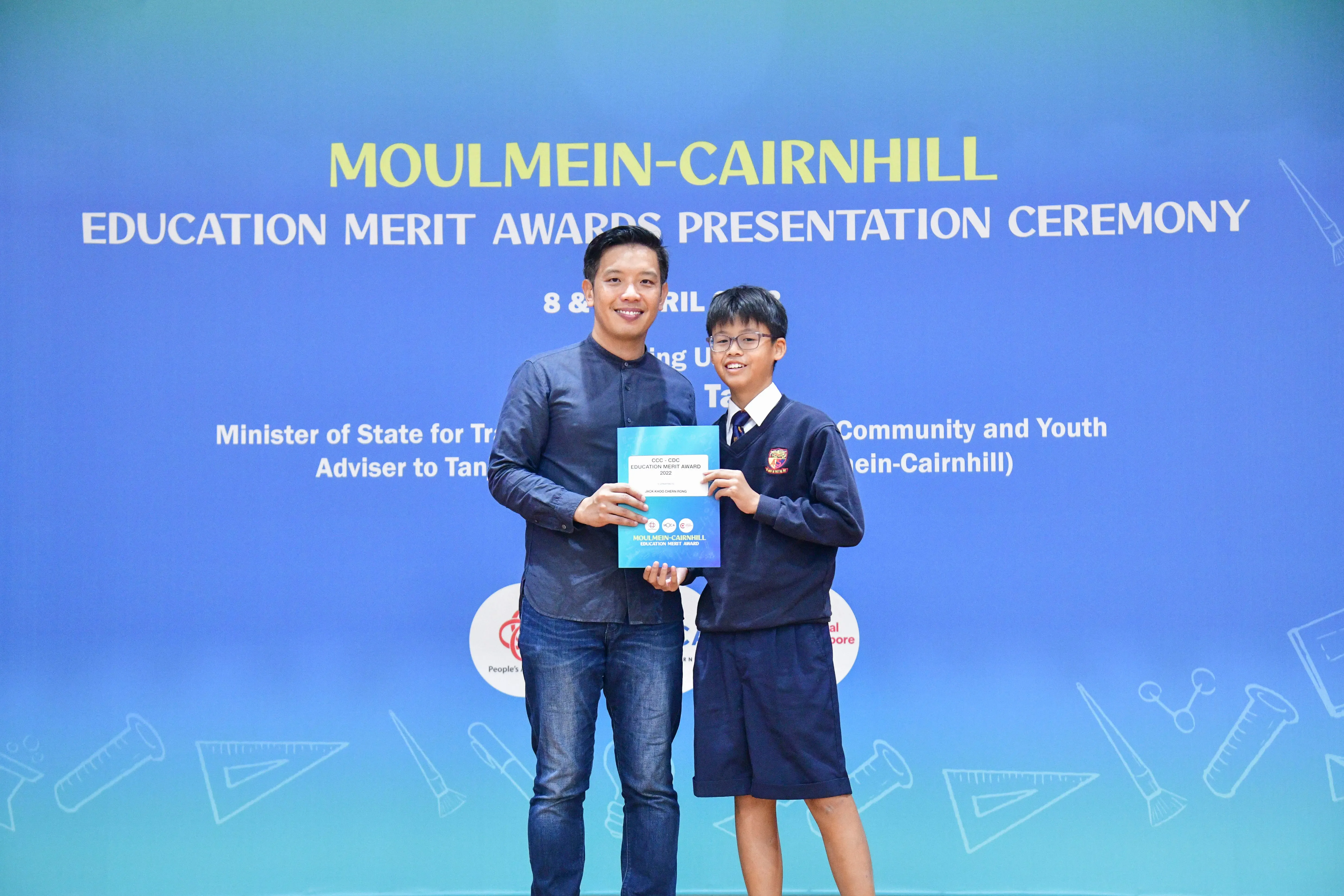 Adult man and young boy in school uniform hold a certificate at Moulmein-Cairnhill Education Merit Awards ceremony.