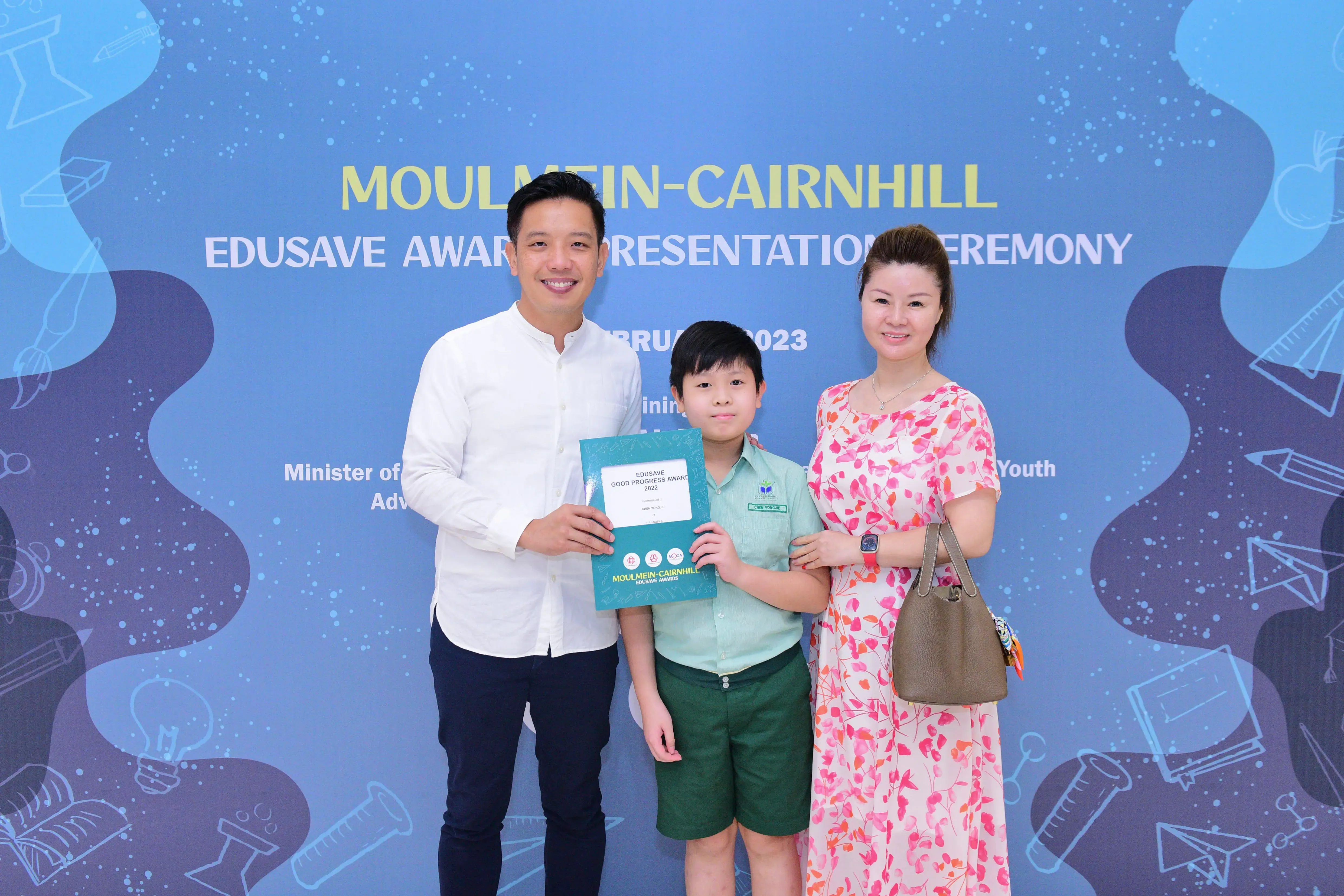 A boy holding an Edusave award certificate stands between two adults in front of a Moulmein-Cairnhill ceremony banner.
