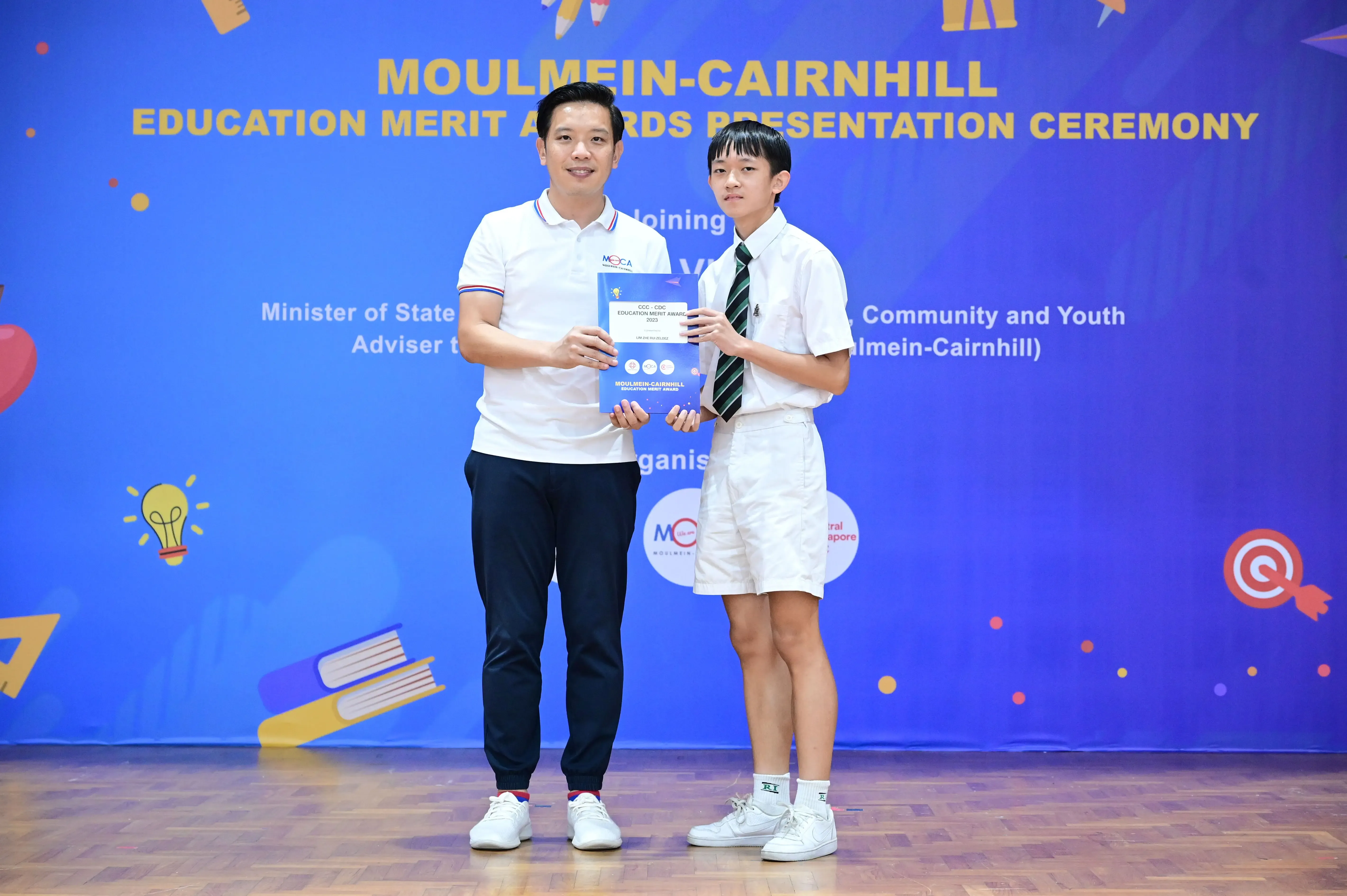 An adult and a student in school uniform hold a certificate at the Moulmein-Cairnhill Education Merit Awards ceremony.