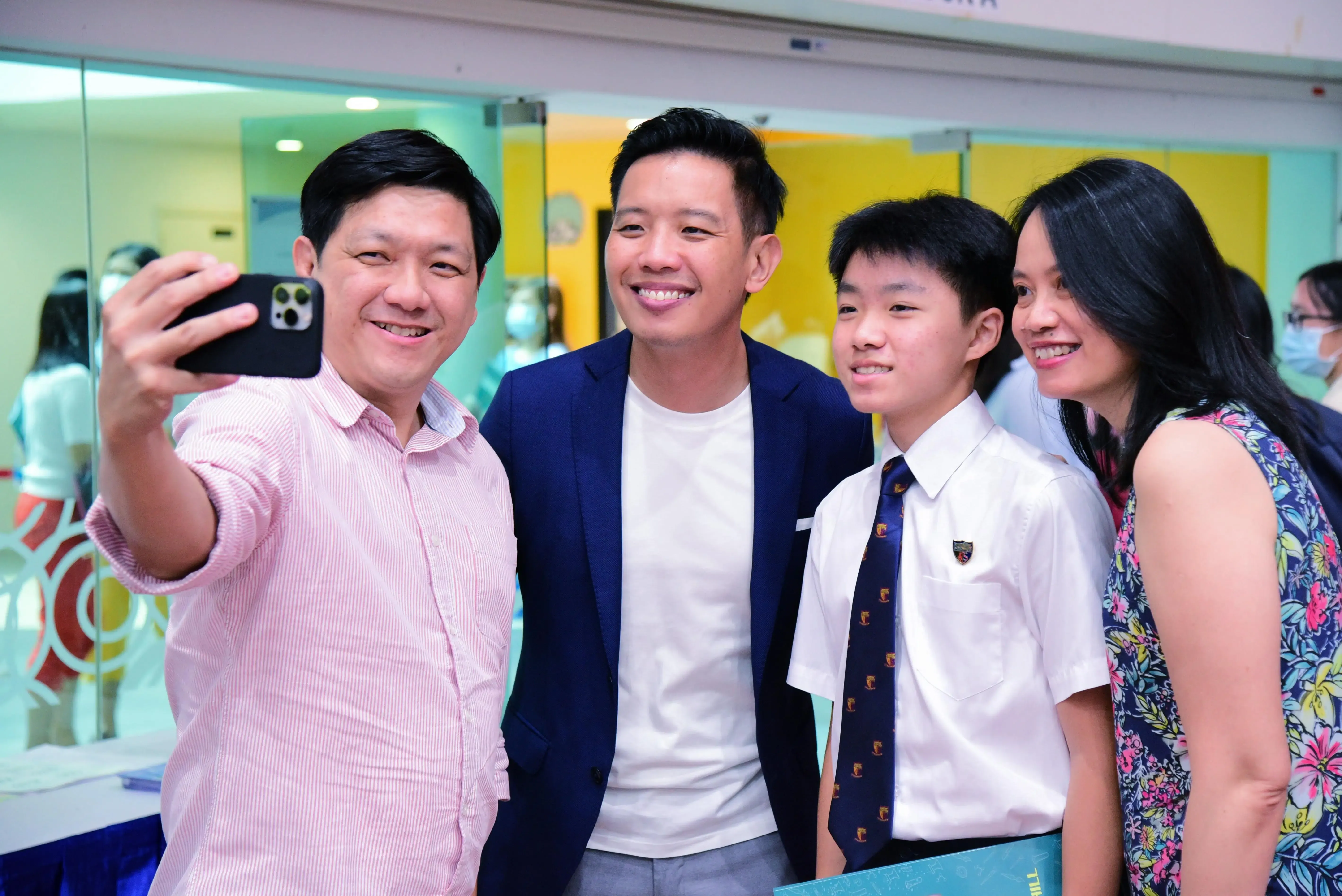 Four smiling people pose for a selfie, including a young student in uniform and tie, indoors.