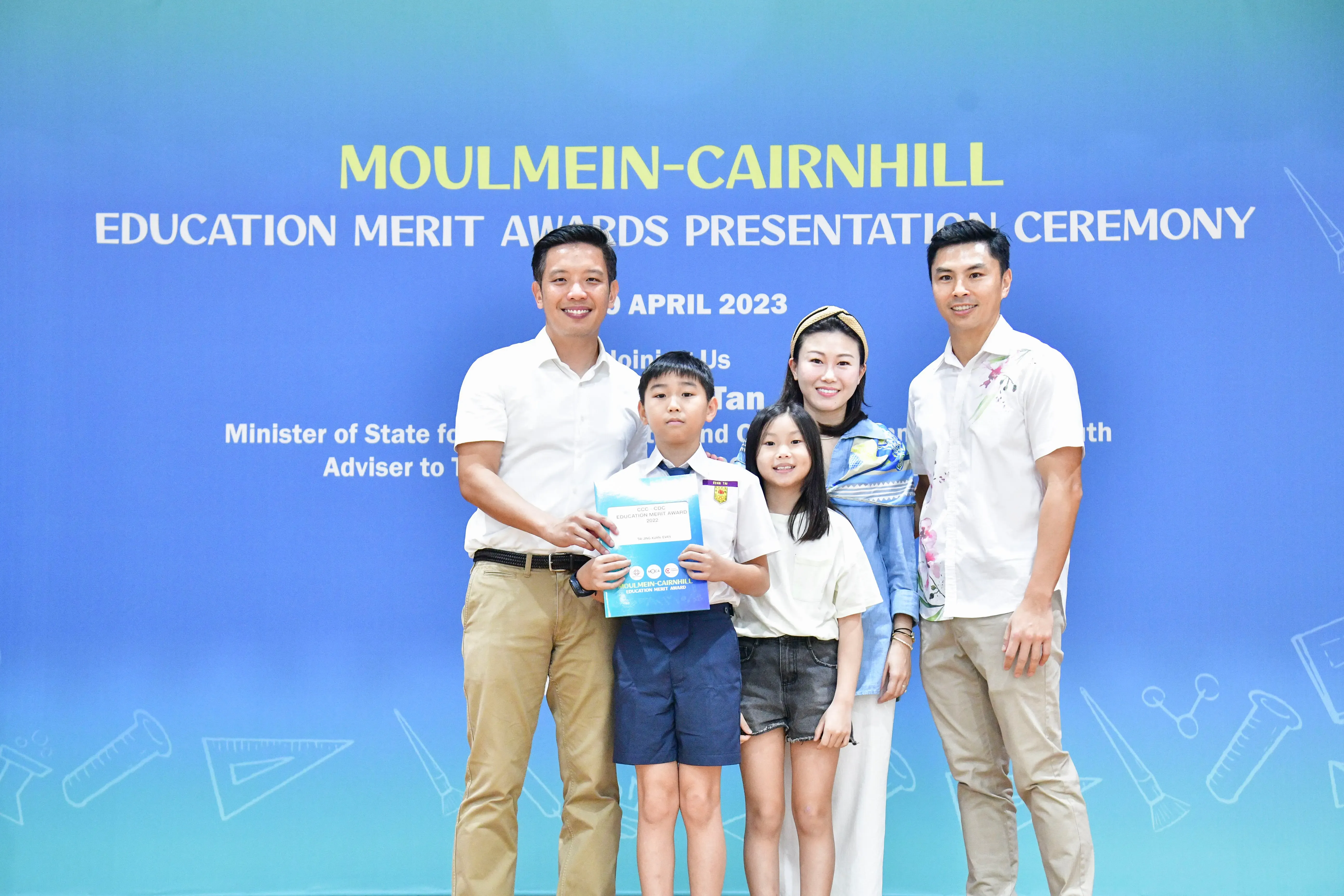 Five people pose before a Moulmein-Cairnhill Education Merit Awards Presentation Ceremony banner in April 2023.
