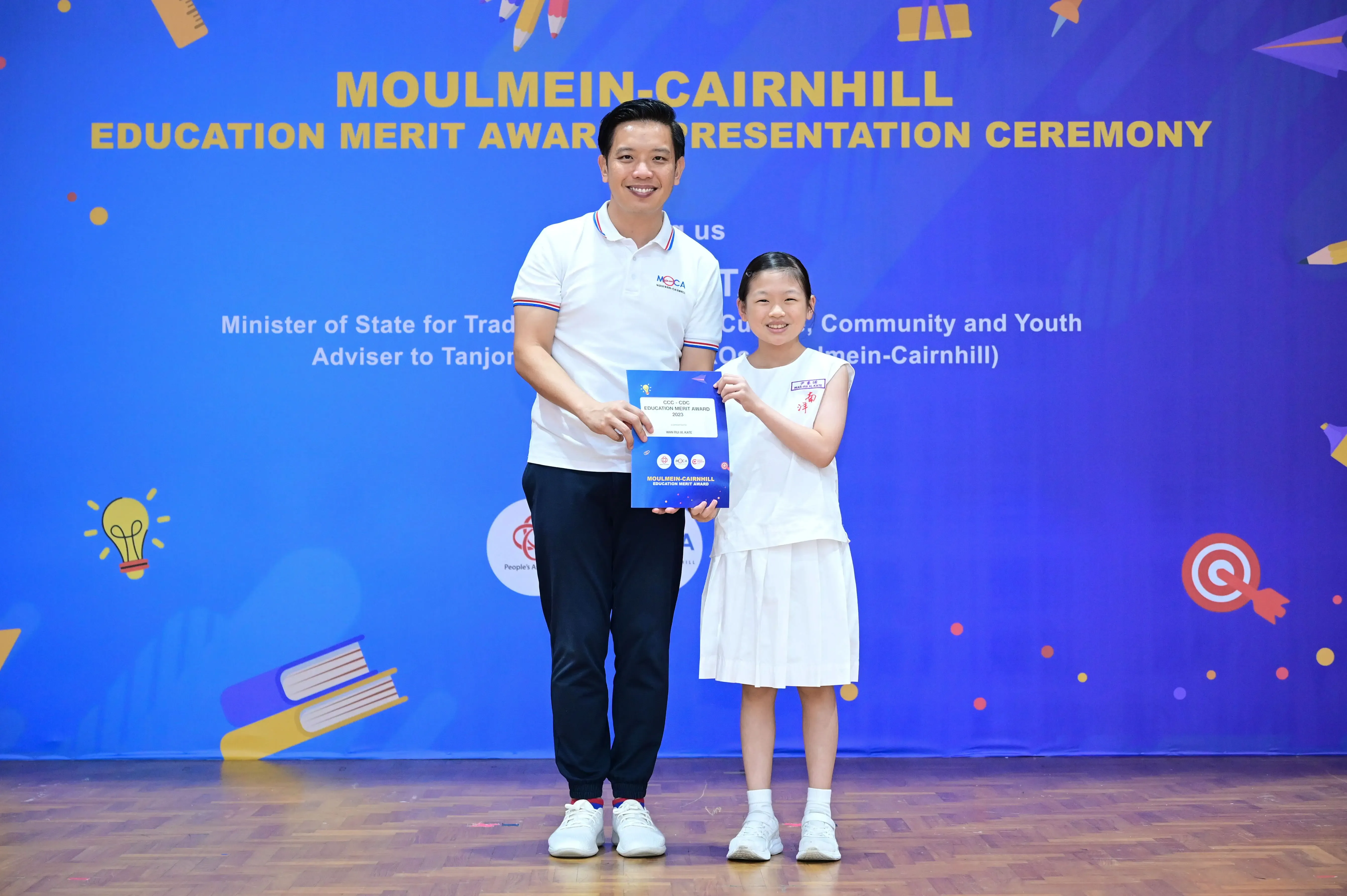 A man and a young girl hold a blue award certificate at the Moulmein-Cairnhill Education Merit Award Ceremony.