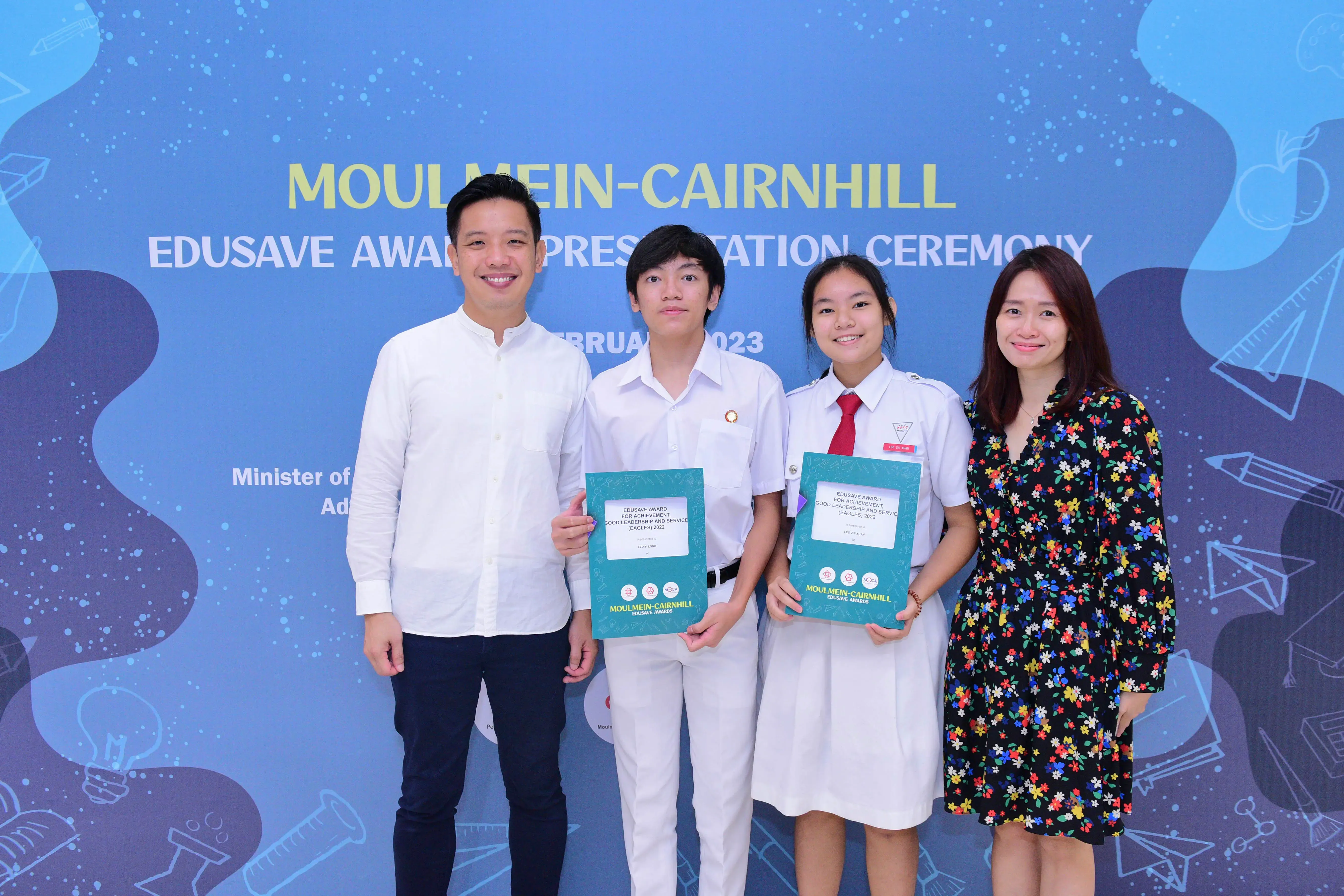 Four people pose in front of a Moulmein-Cairnhill Edusave Award Presentation Ceremony banner, two students holding certificates.