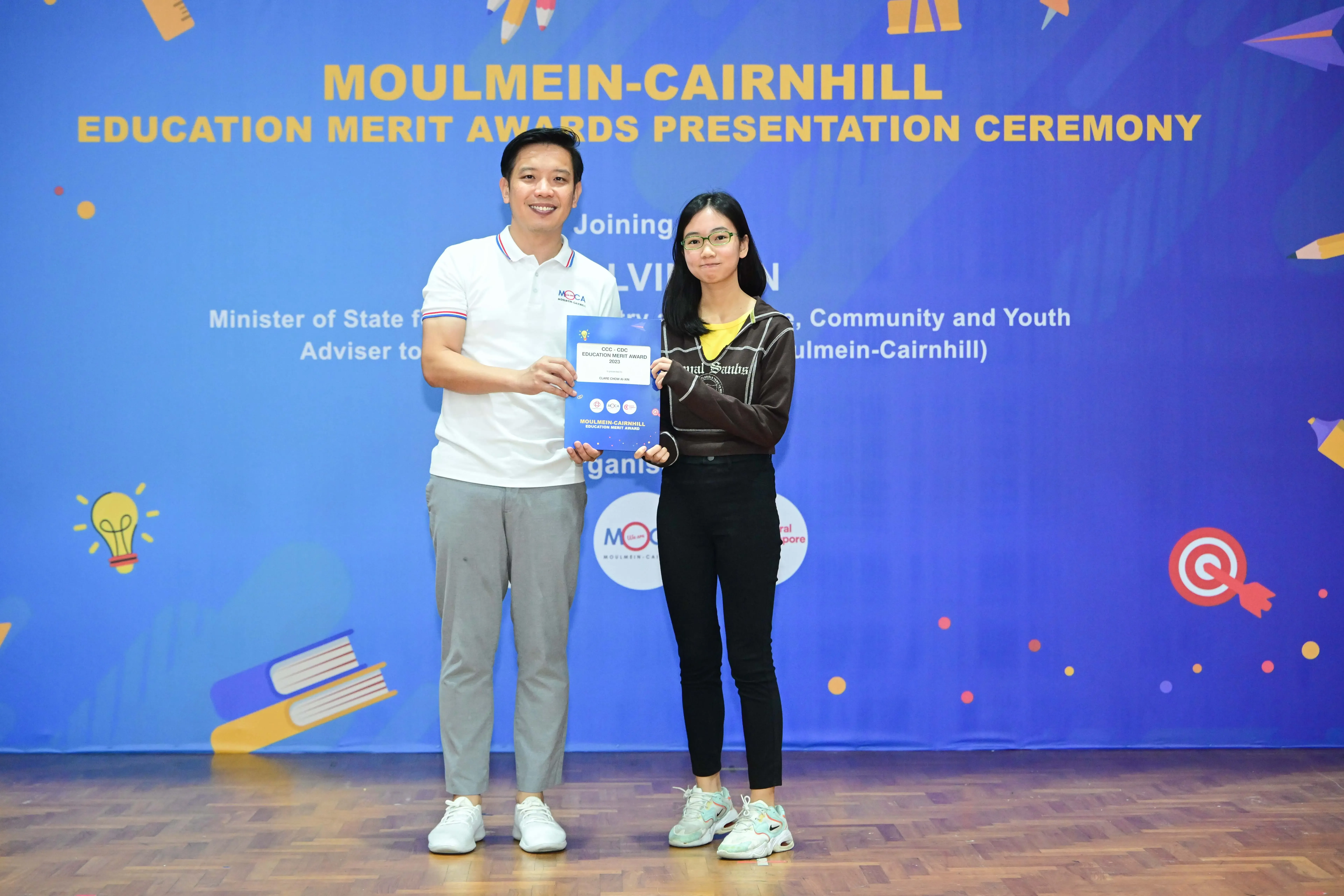 Two people stand holding an award booklet at the Moulmein-Cairnhill Education Merit Awards Presentation Ceremony.