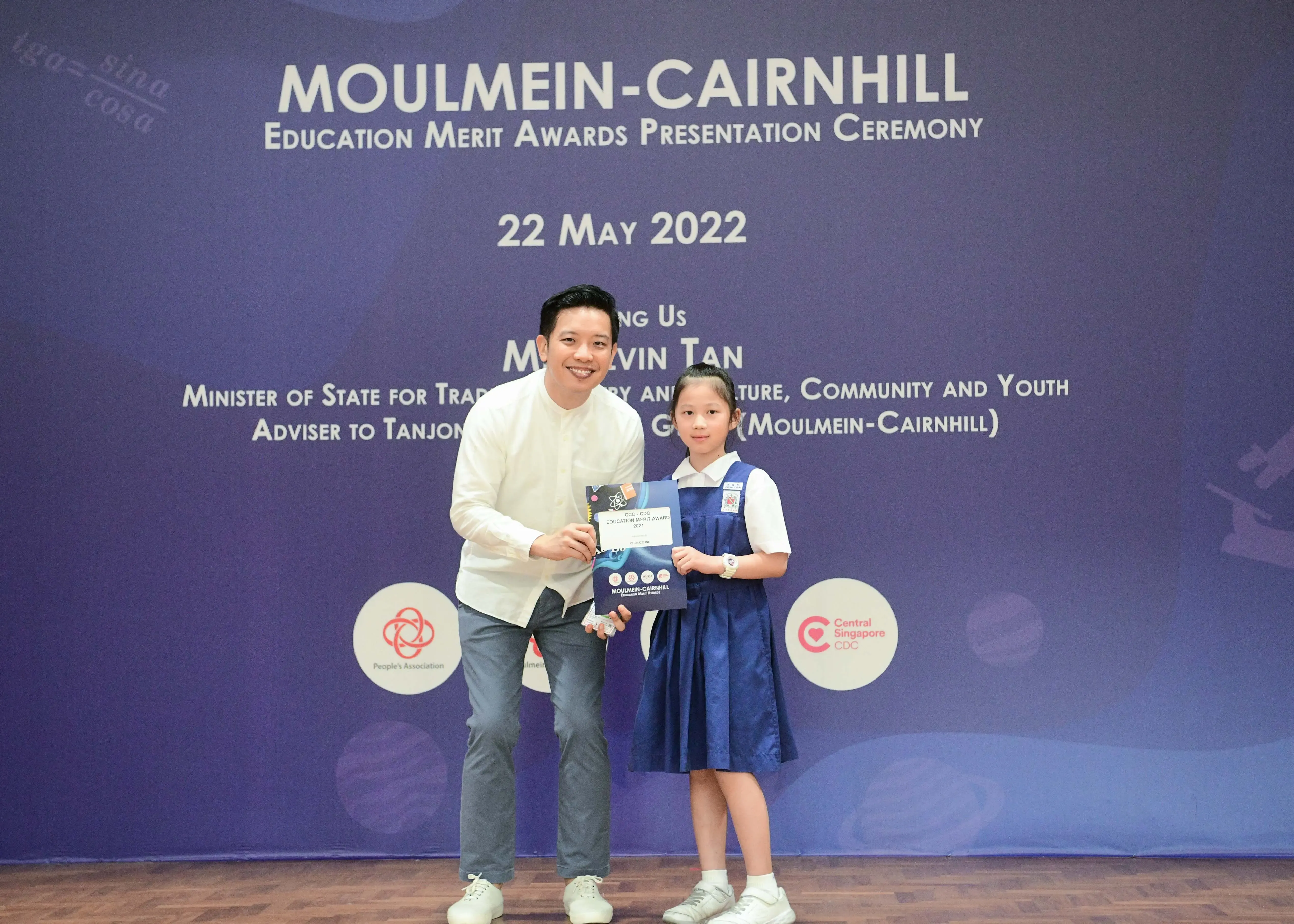A man and a young girl in school uniform hold an award certificate at the Moulmein-Cairnhill Education Merit Awards ceremony.