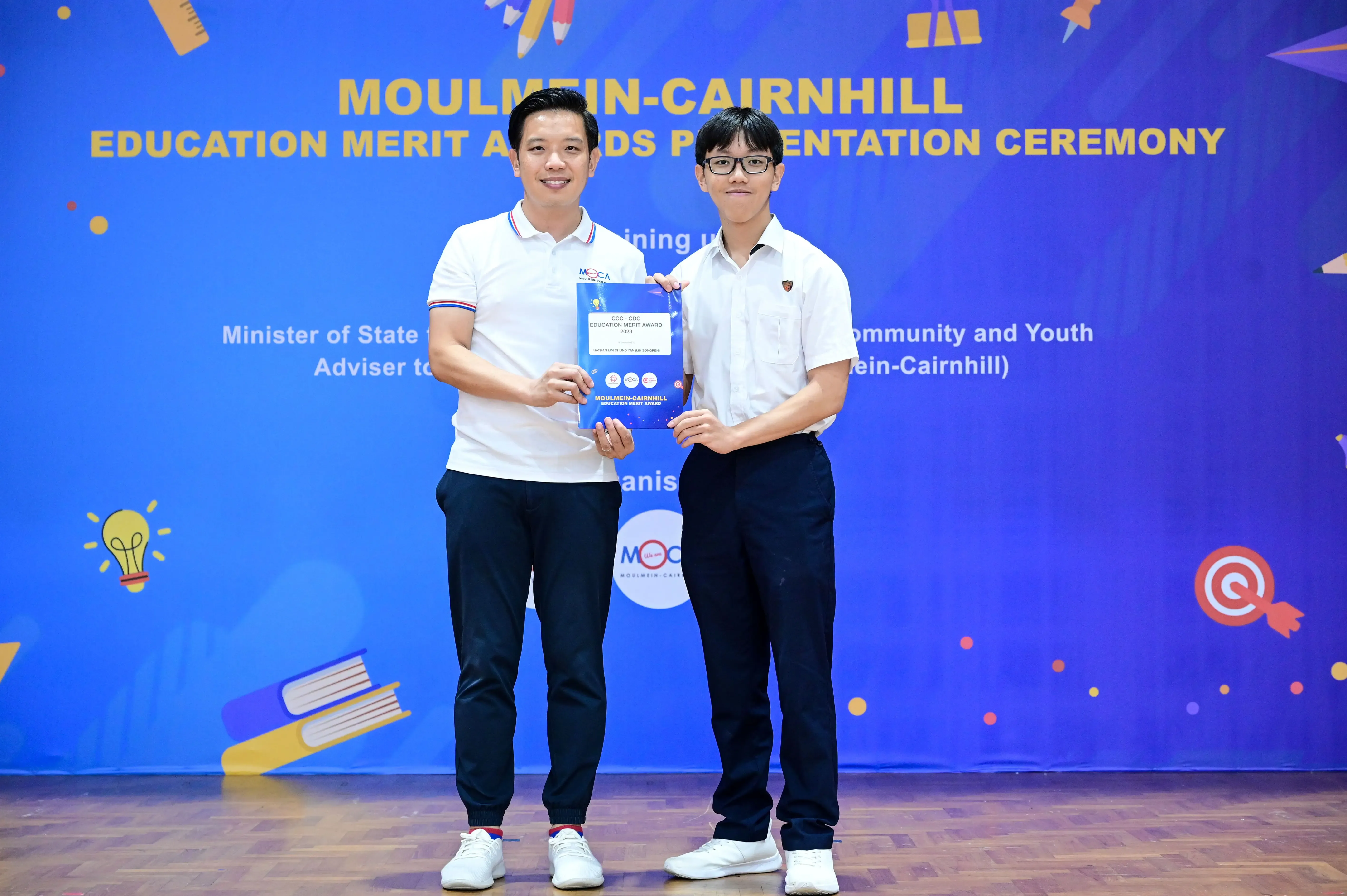 Two men hold an award booklet at the Moulmein-Cairnhill Education Merit Awards Presentation Ceremony.