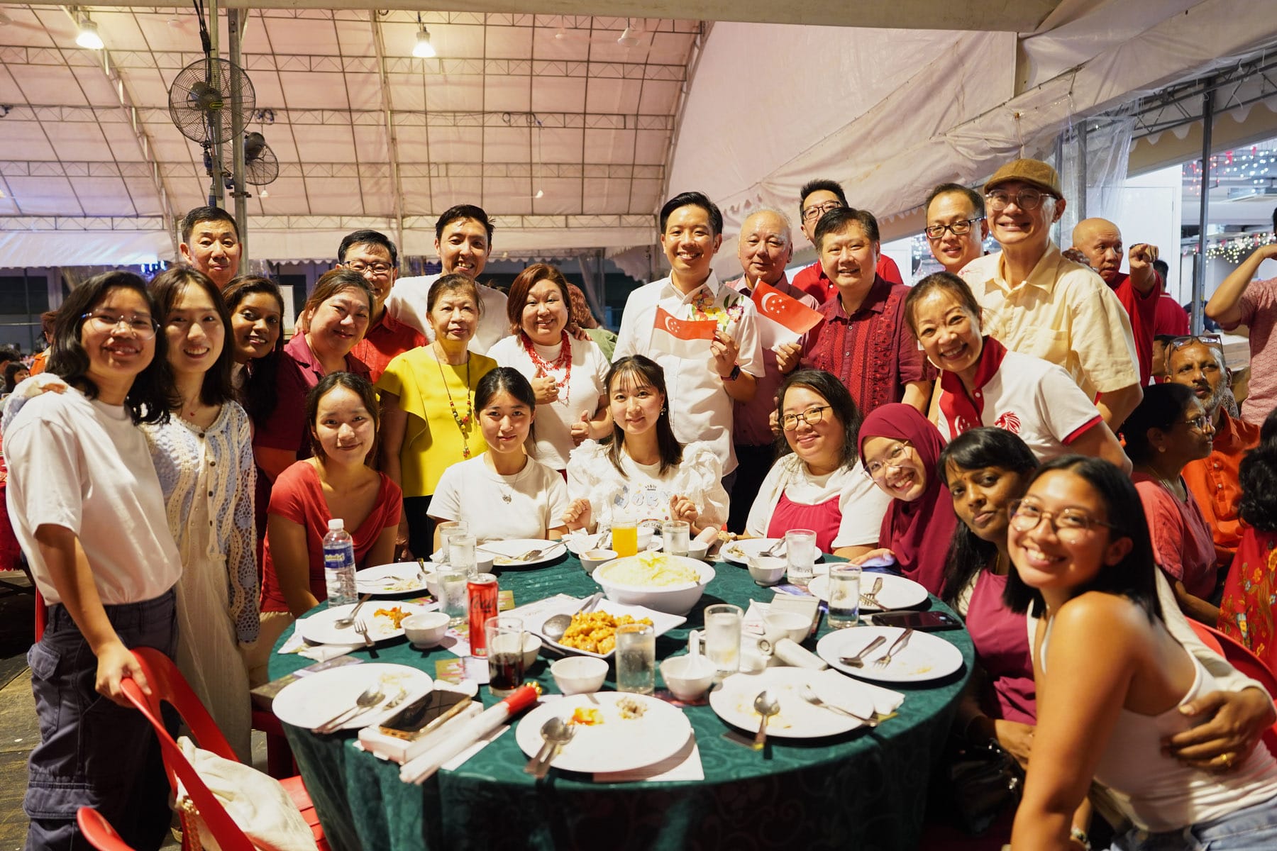A large group of smiling people pose together at a round dining table under a tent, with Singapore flags visible.