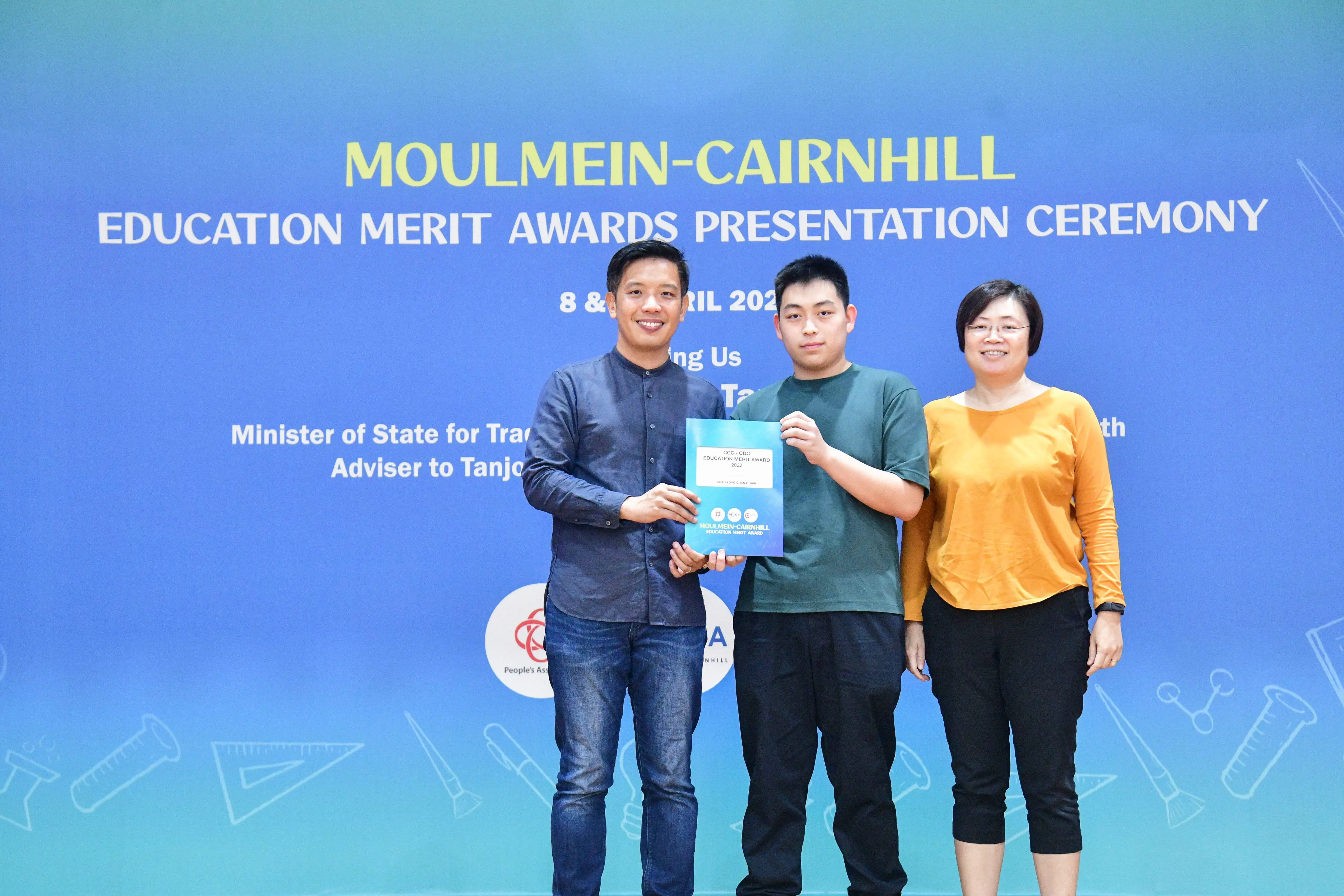 Three people stand before a blue banner for the Moulmein-Cairnhill Education Merit Awards Presentation Ceremony, holding a certificate.