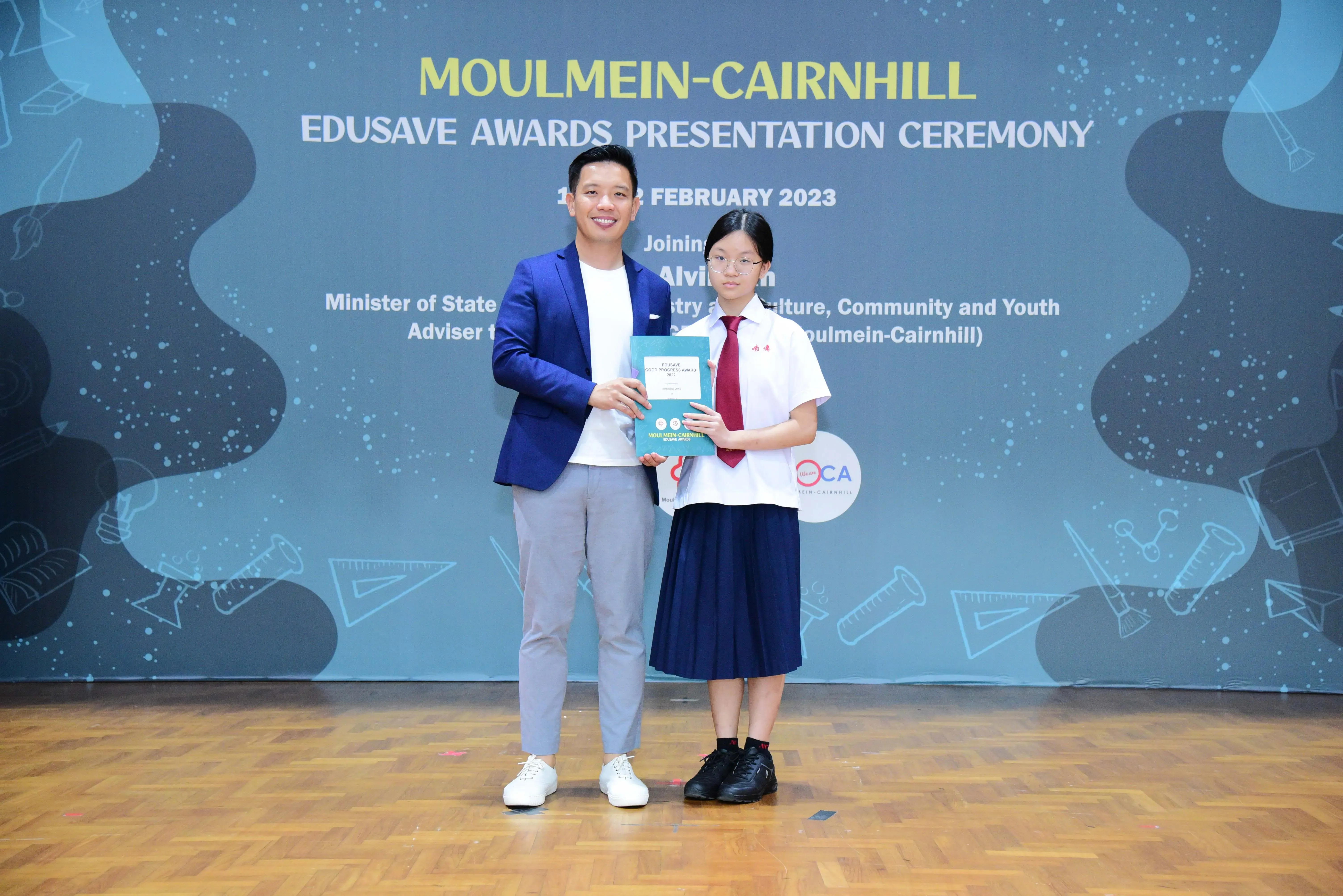 A man in a blue blazer and a young girl in school uniform hold an award at the Moulmein-Cairnhill Edusave Awards Ceremony 2023.