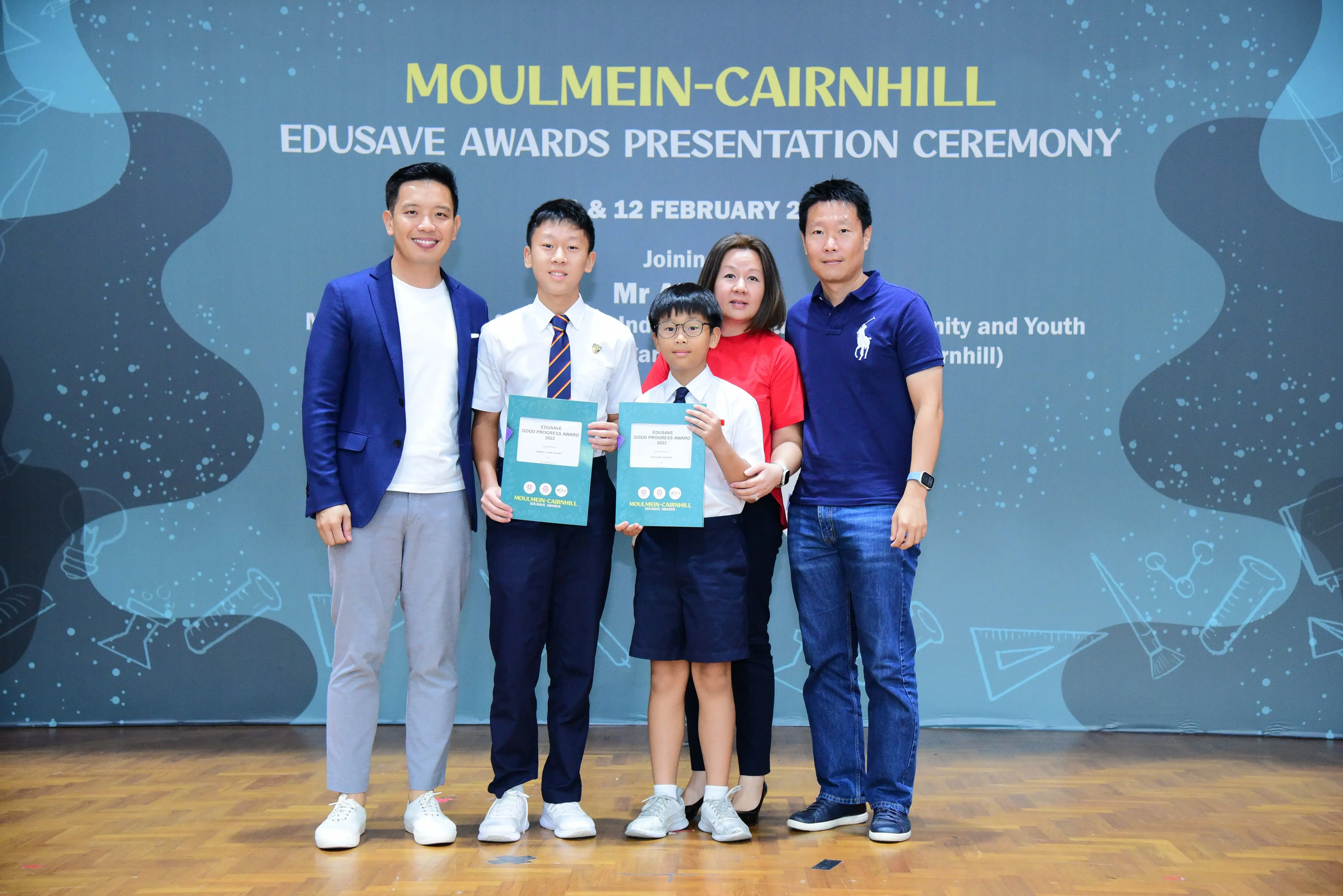 Two boys holding award certificates pose with three adults at the Moulmein-Cairnhill Edusave Awards Presentation Ceremony.
