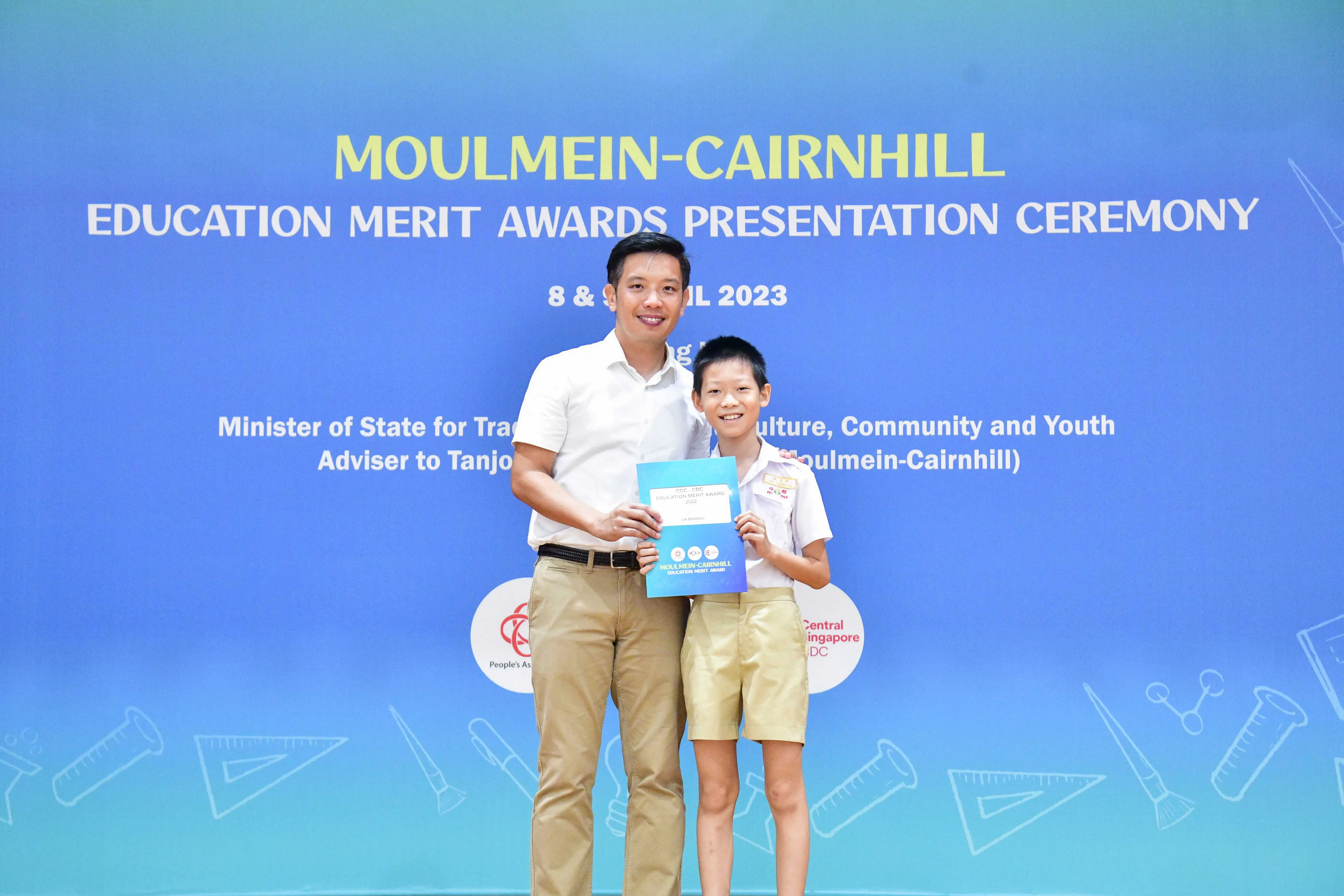 Adult man and young boy holding an award booklet at the Moulmein-Cairnhill Education Merit Awards Ceremony 2023.