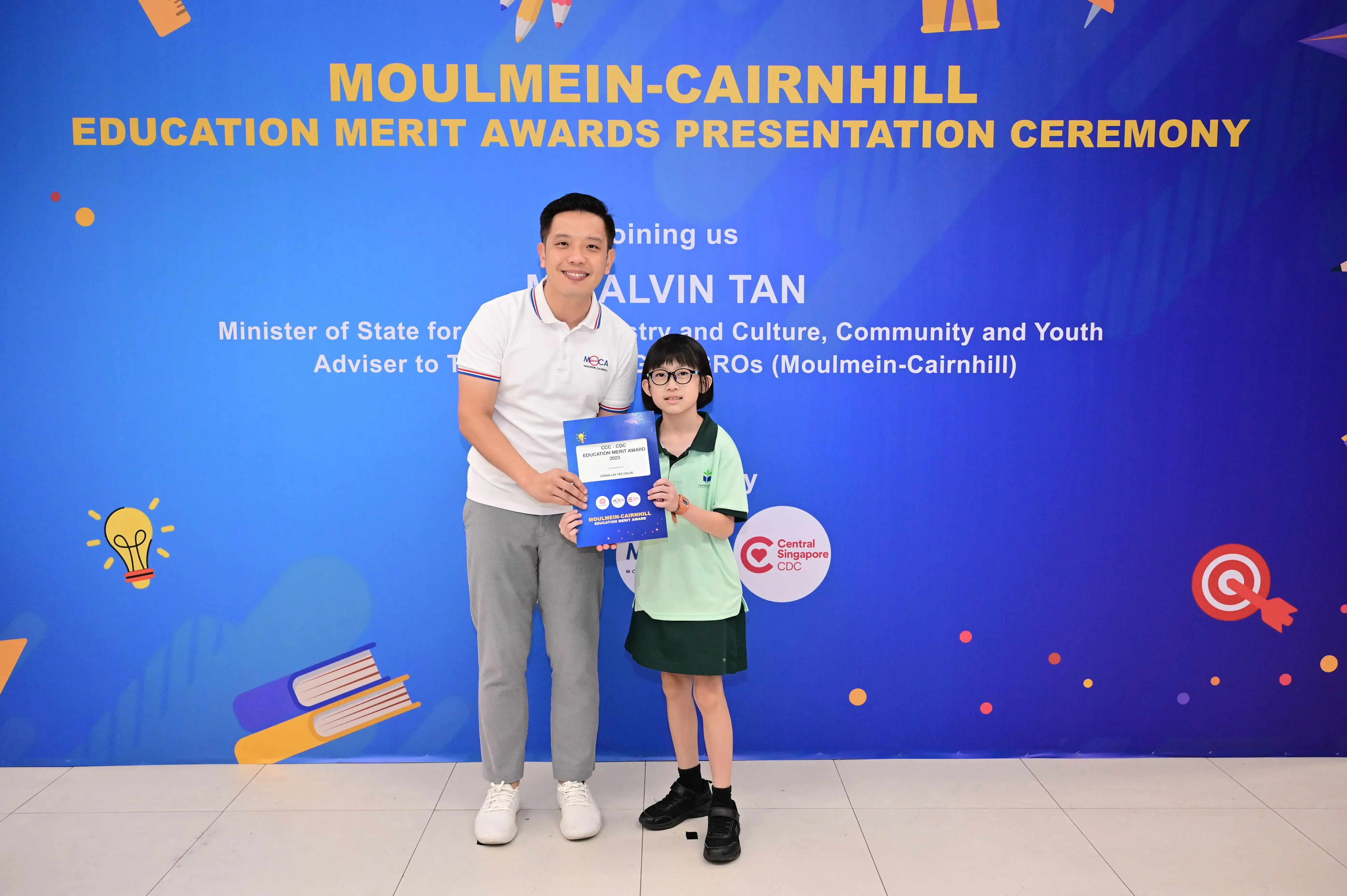 A man and a young girl hold a blue award folder at the Moulmein-Cairnhill Education Merit Awards Presentation Ceremony.