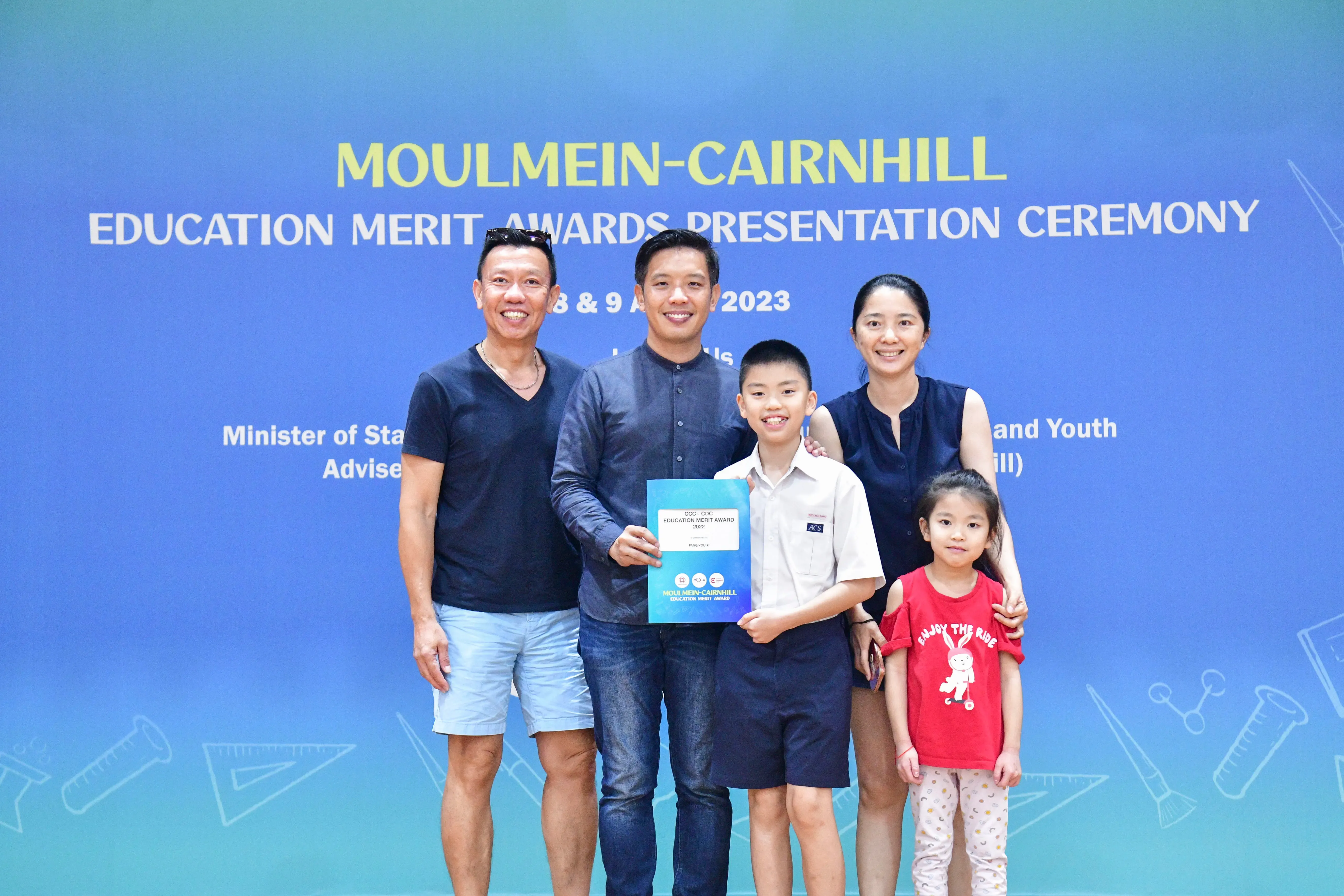 Four adults and two children pose before a Moulmein-Cairnhill Education Merit Awards Presentation Ceremony banner in 2023.