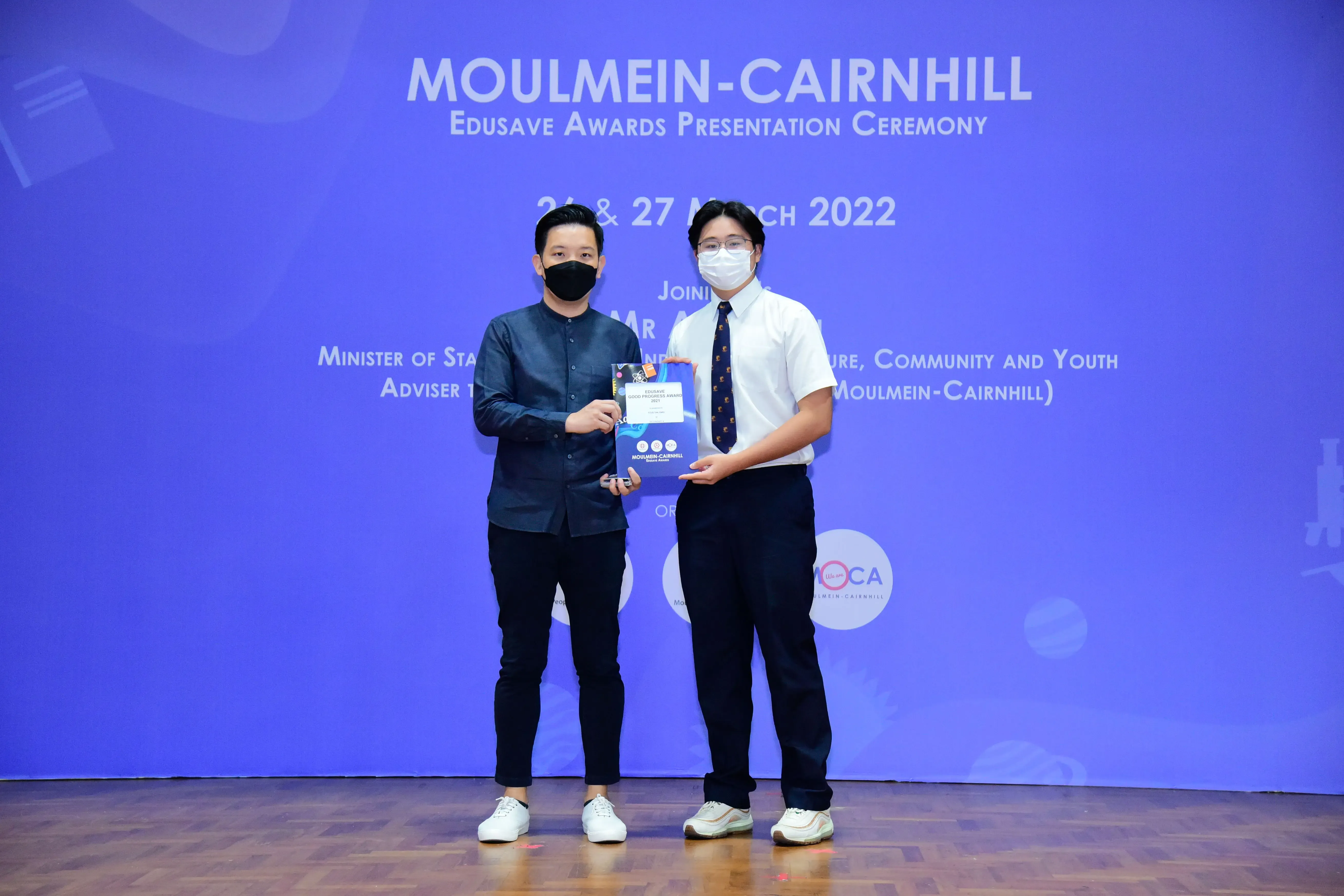 Two masked men hold an award booklet on stage at the Moulmein-Cairnhill Edusave Awards Ceremony, March 2022.