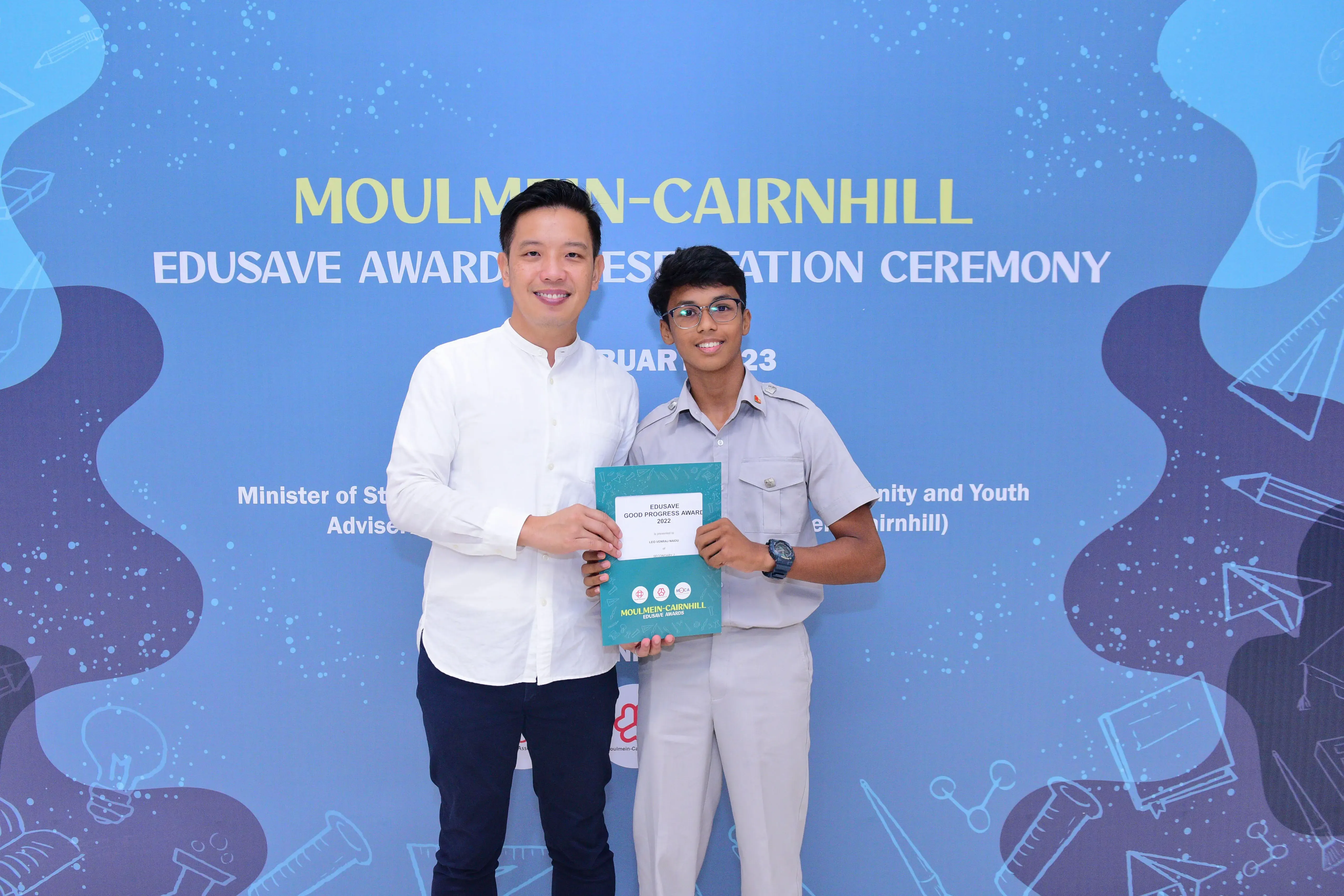 Two men smile holding a teal Edusave Good Progress Award certificate at the Moulmein-Cairnhill ceremony backdrop.