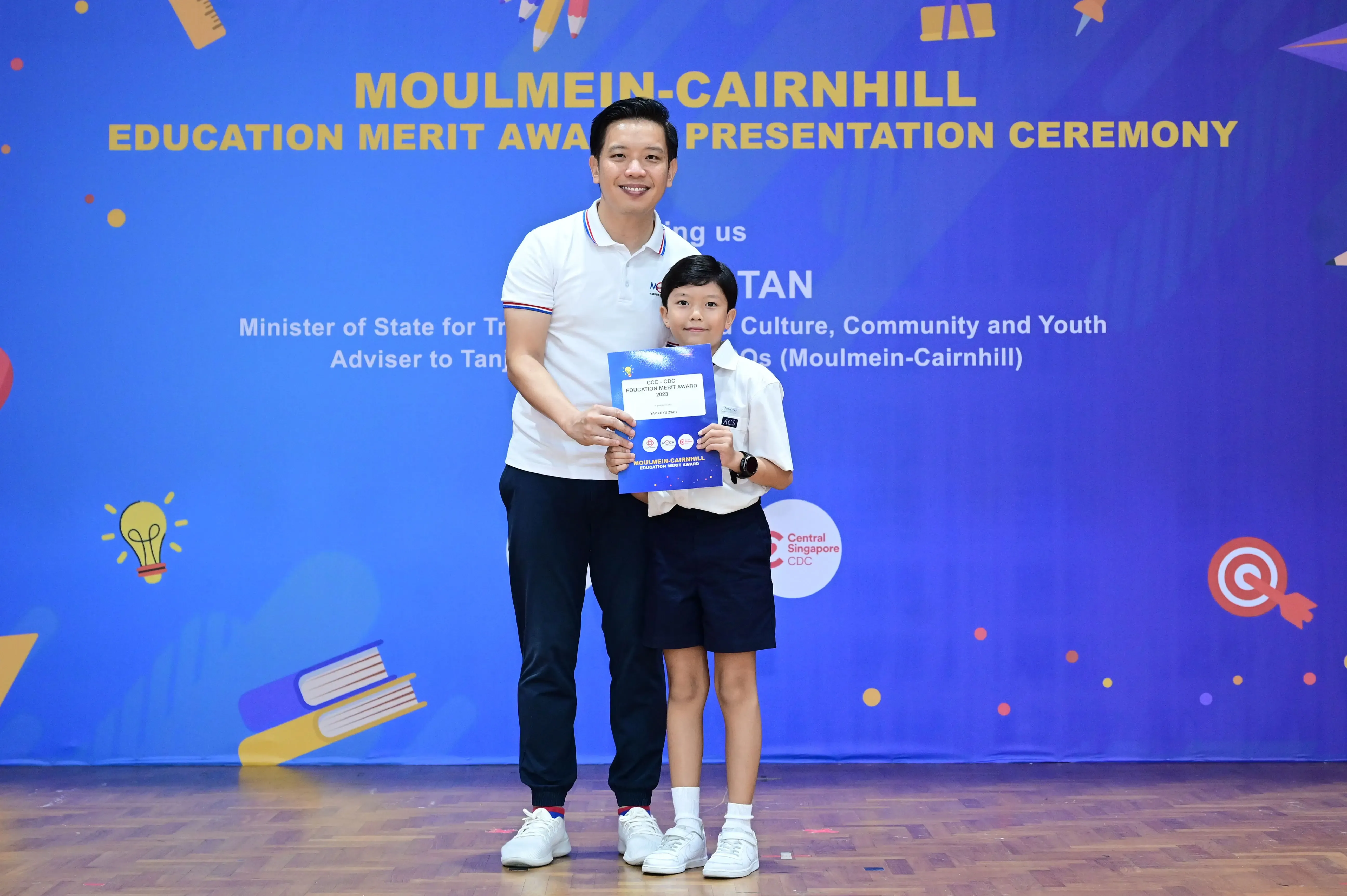 A man and a young boy hold a blue award certificate at the Moulmein-Cairnhill Education Merit Award Ceremony.