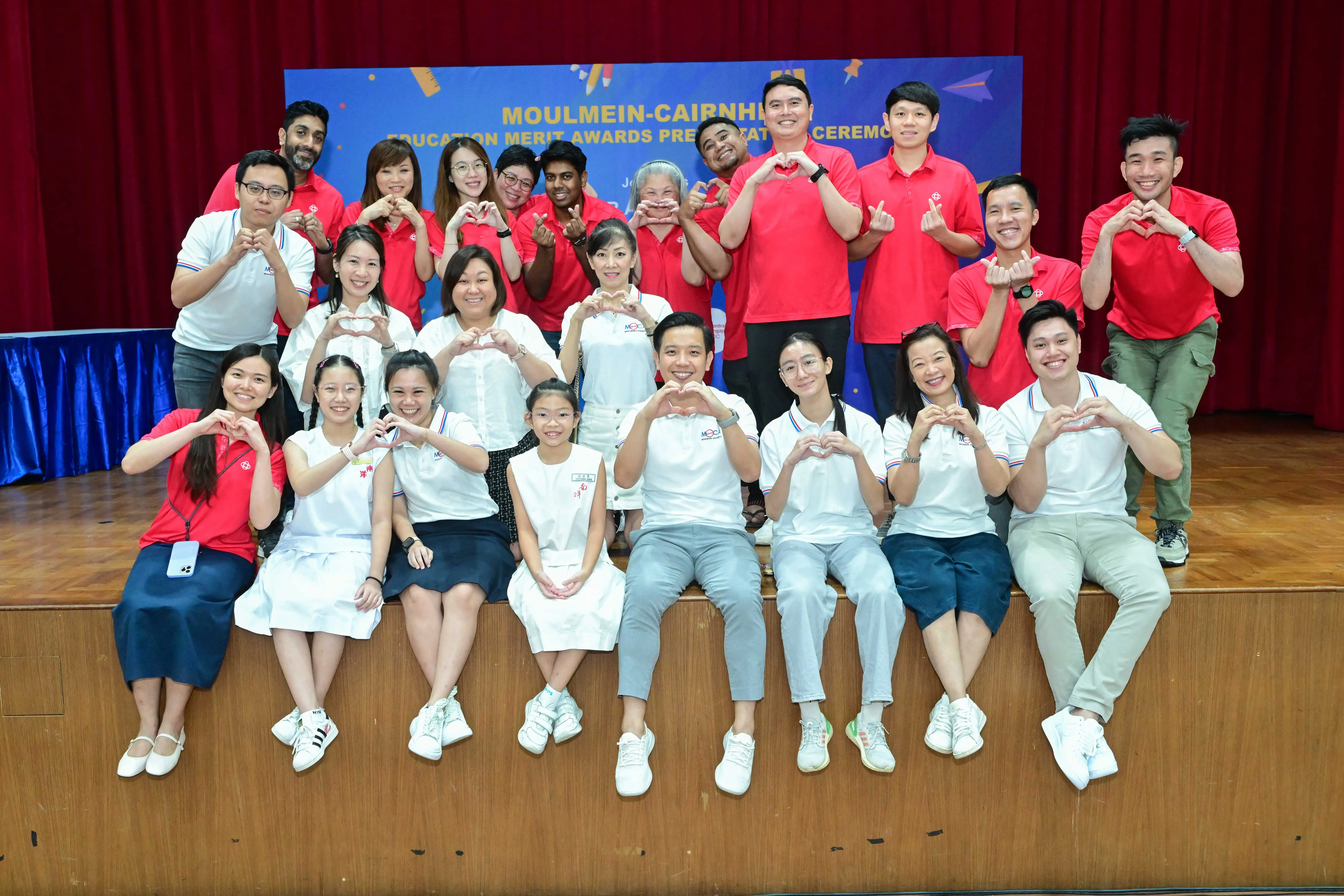 Group of people in red and white shirts posing with heart hand gestures at Moulmein-Cairnhill Education Merit Awards ceremony.