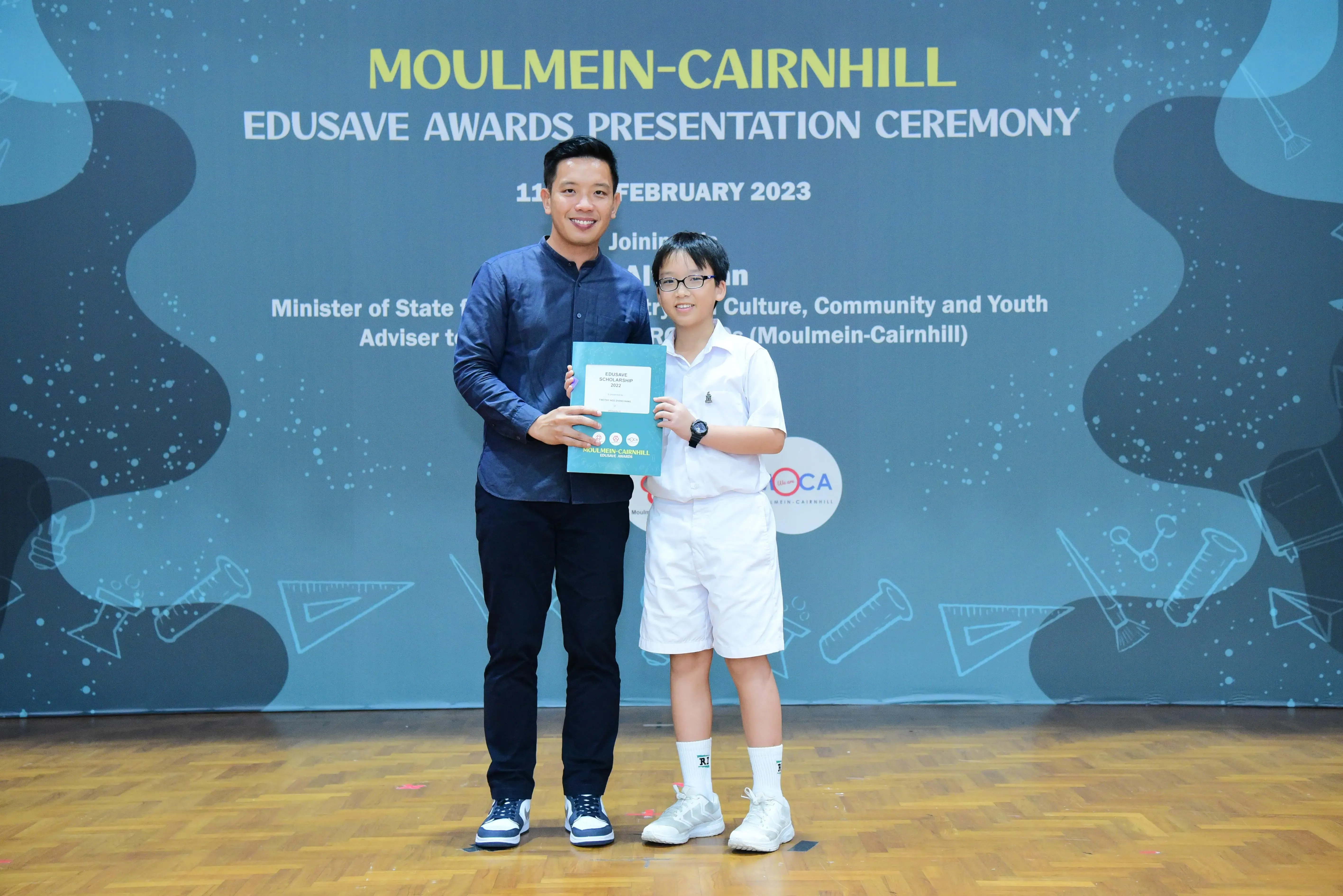 An adult and a young student hold a certificate at the Moulmein-Cairnhill Edusave Awards Presentation Ceremony 2023.