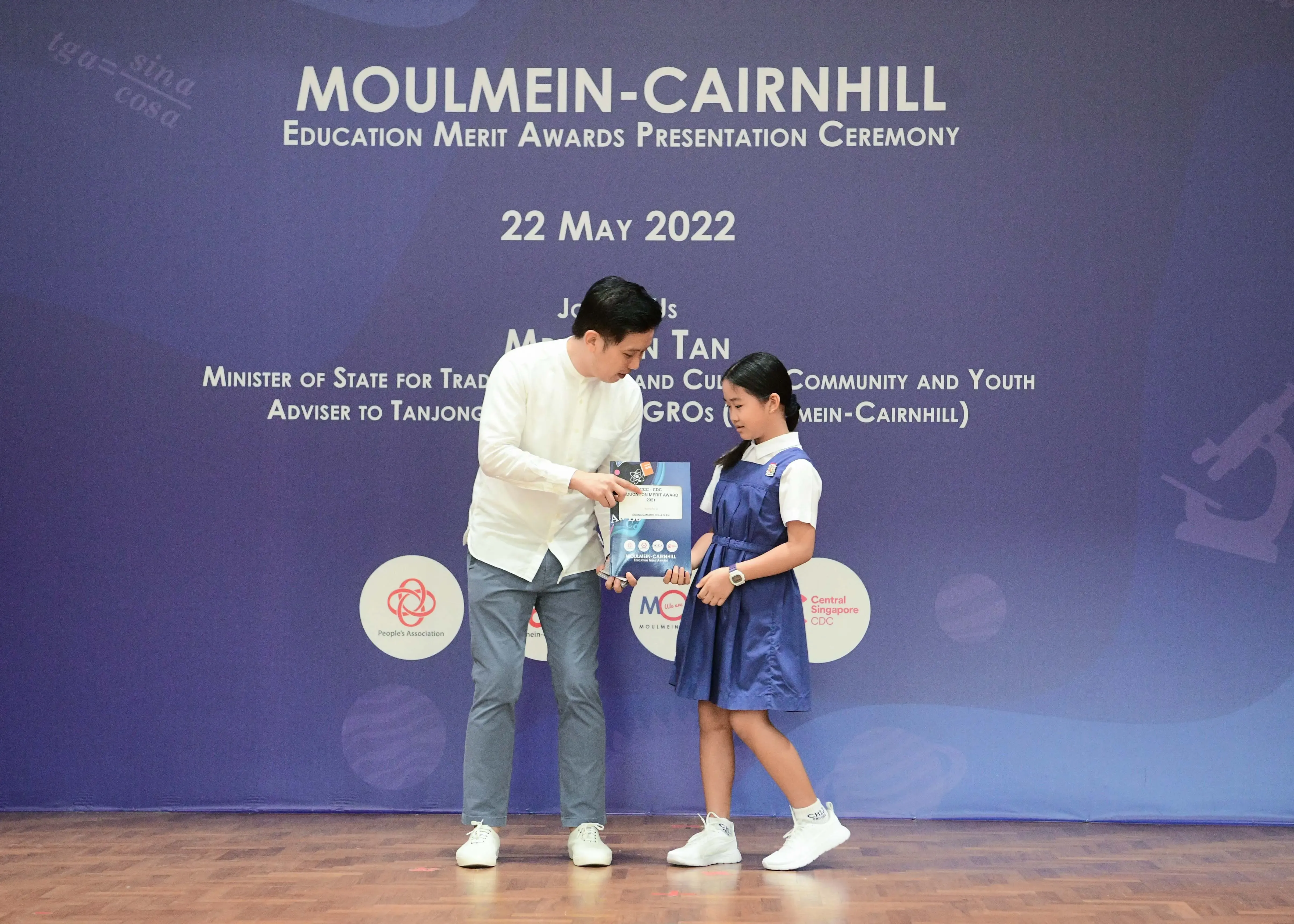A man presents an award to a young girl in a blue school uniform at the Moulmein-Cairnhill Education Merit Awards, May 2022.