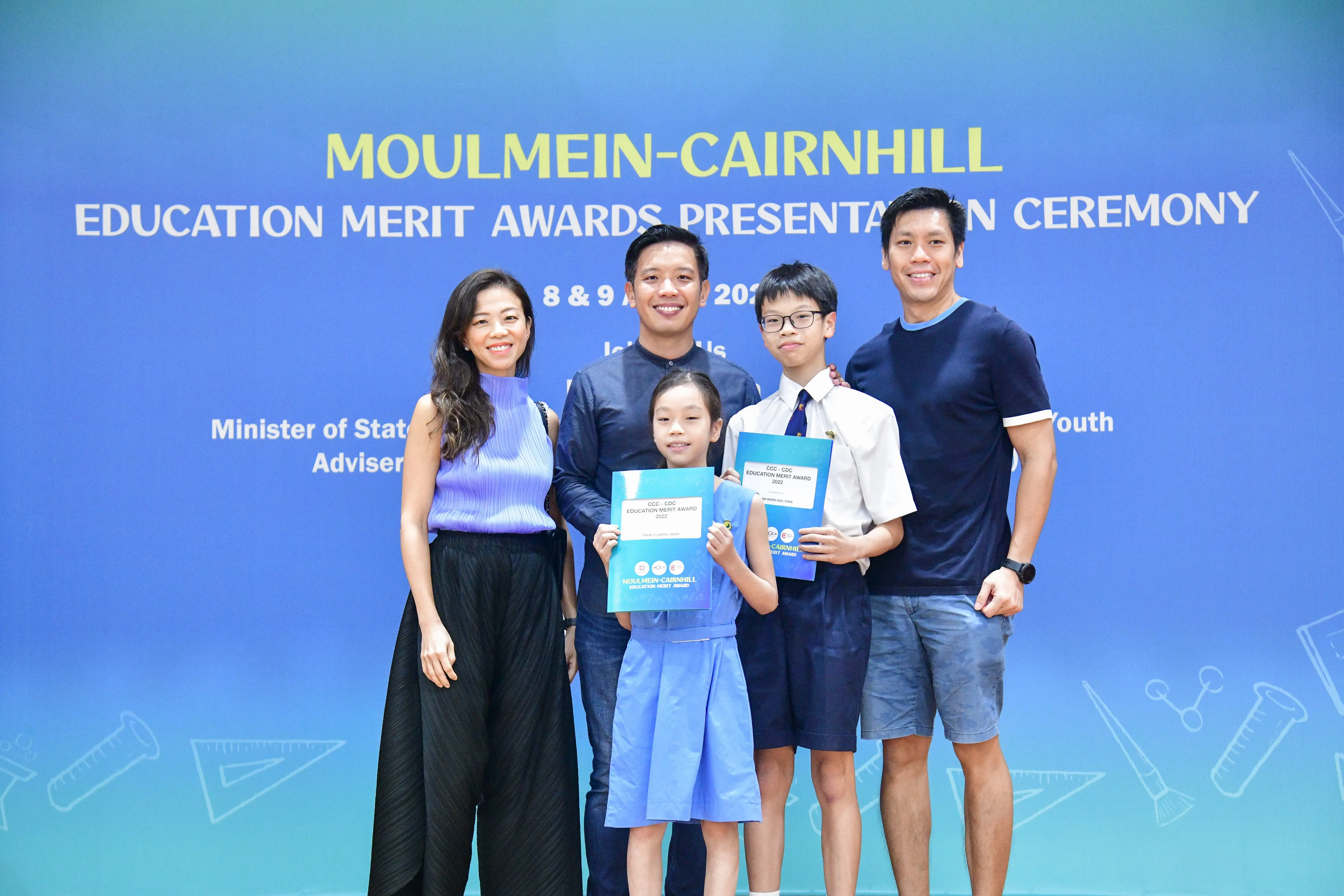 Five people pose before a Moulmein-Cairnhill Education Merit Awards banner, two children holding award certificates.