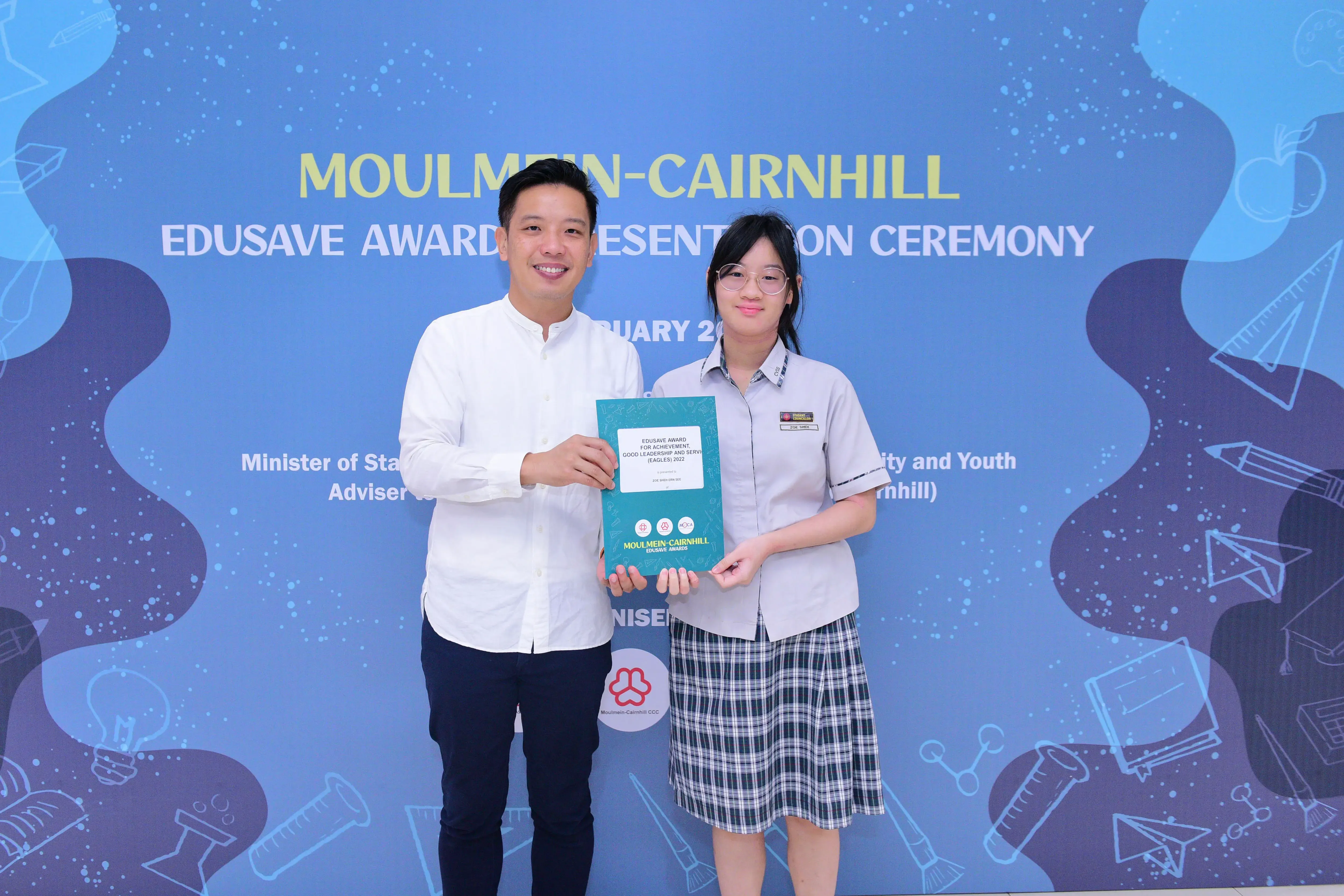 Two people hold a certificate together in front of a Moulmein-Cairnhill Edusave Awards Presentation Ceremony backdrop.